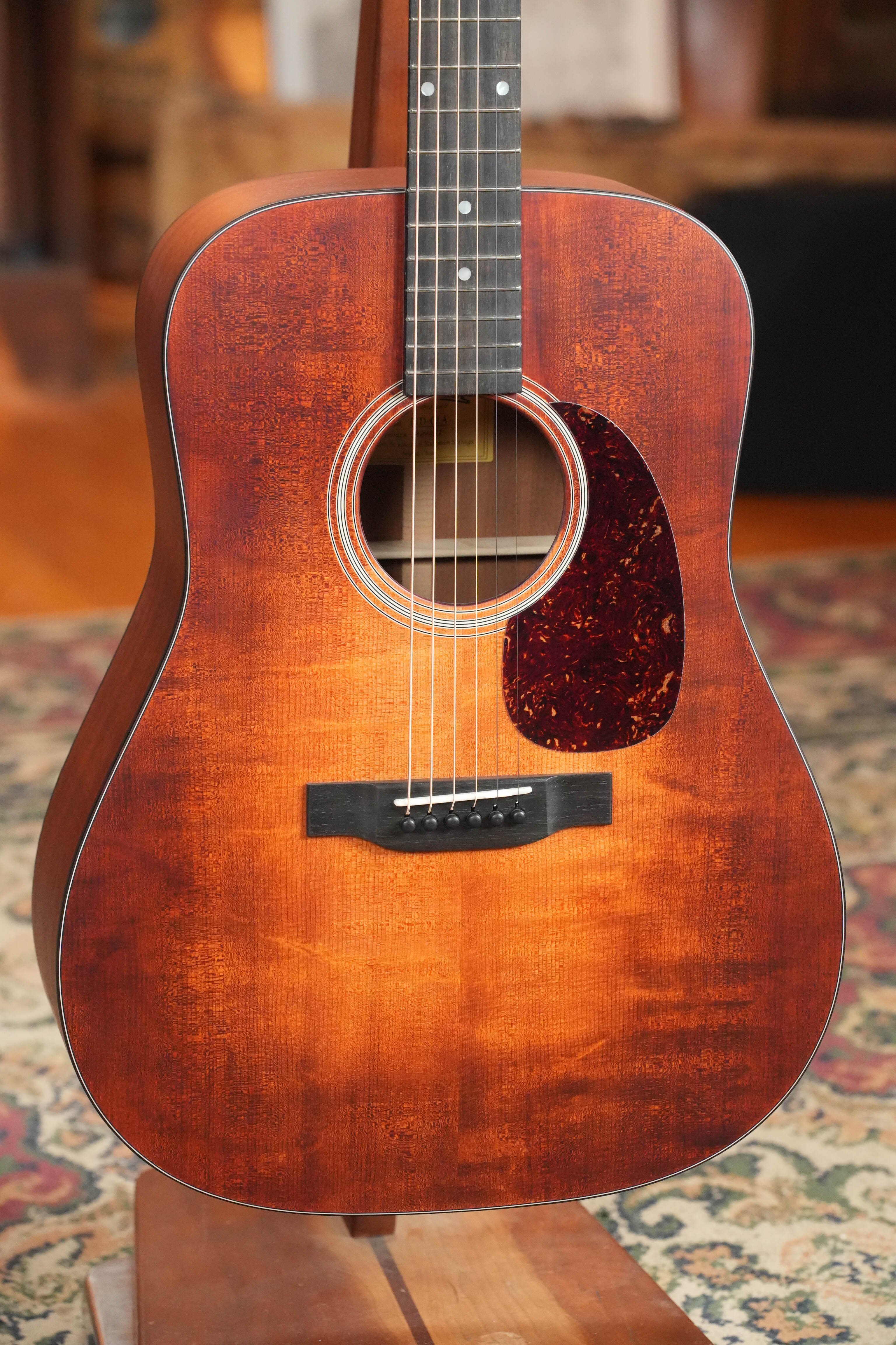 A close-up of the Eastman Guitars E1D-CLA Classic Finish Dreadnought Acoustic Guitar #1172 with a tortoiseshell pickguard, resting on a wooden stand over a patterned rug. The solid Sitka spruce top has a rustic, worn texture.