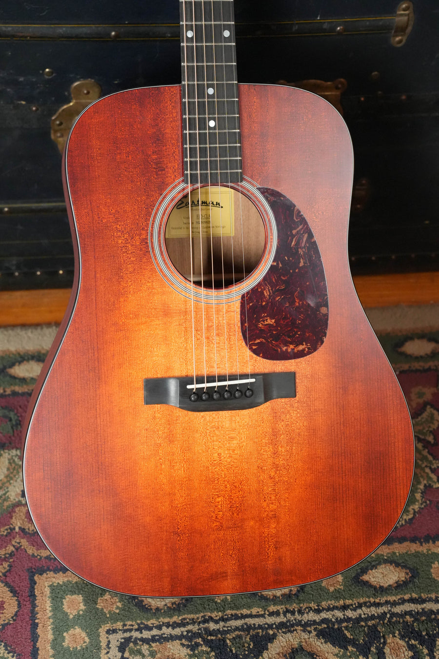 The Eastman Guitars E1D-CLA Classic Finish Dreadnought Acoustic Guitar #9429 with solid Sitka spruce top and dark pickguard stands upright on a patterned rug, a vintage trunk visible in the background.