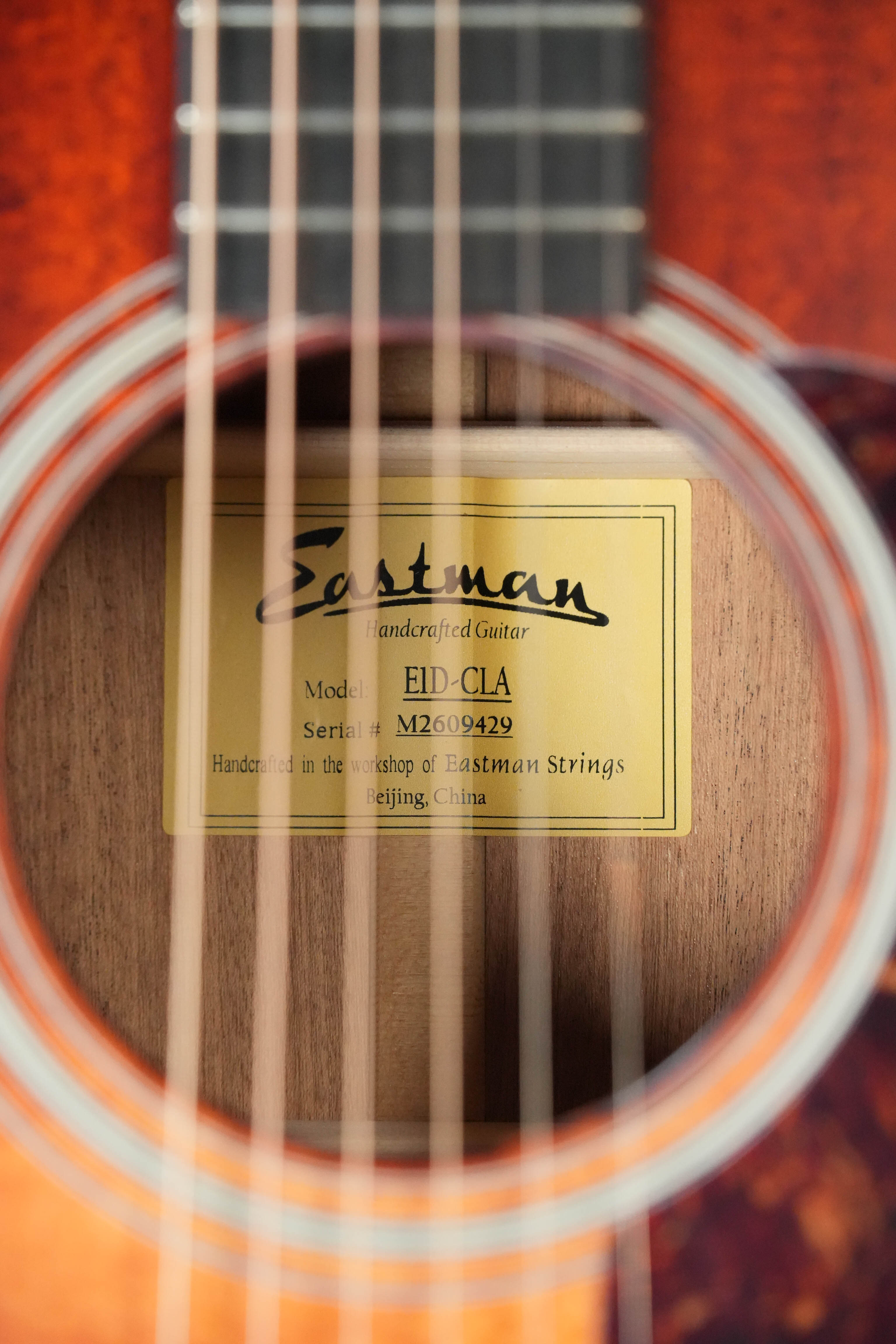 A close-up view through the strings of an Eastman Guitars E1D-CLA Classic Finish Dreadnought Acoustic Guitar #9429 reveals a gold label inside noting its handmade Beijing origin and solid Sitka spruce top.