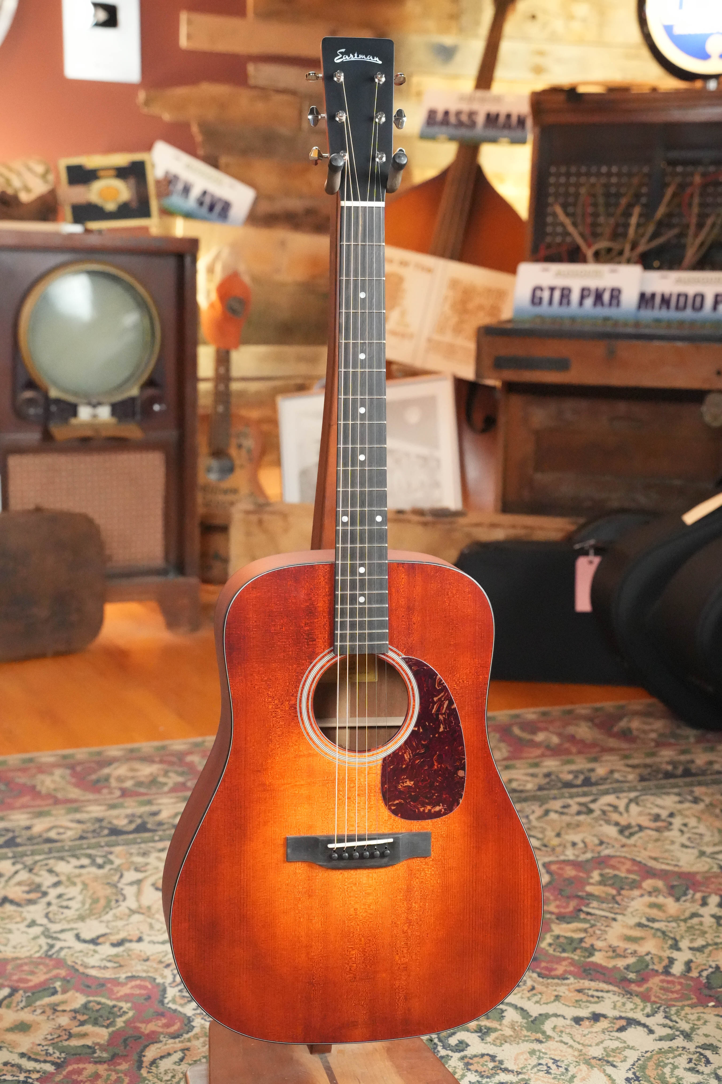 An Eastman Guitars E1D-CLA Classic Finish Dreadnought Acoustic Guitar #9429 stands upright on a patterned rug in a cozy room decorated with vintage musical equipment and rustic furnishings.