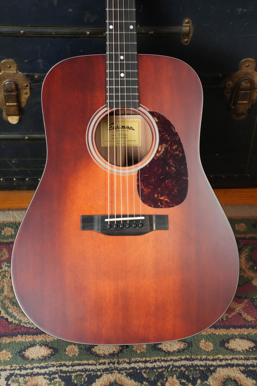 An Eastman E1D-CLA Classic Finish Dreadnought Acoustic Guitar by Eastman Guitars, featuring a tortoiseshell pickguard and solid Sitka spruce top, rests on a patterned rug before a black trunk with brass hardware.