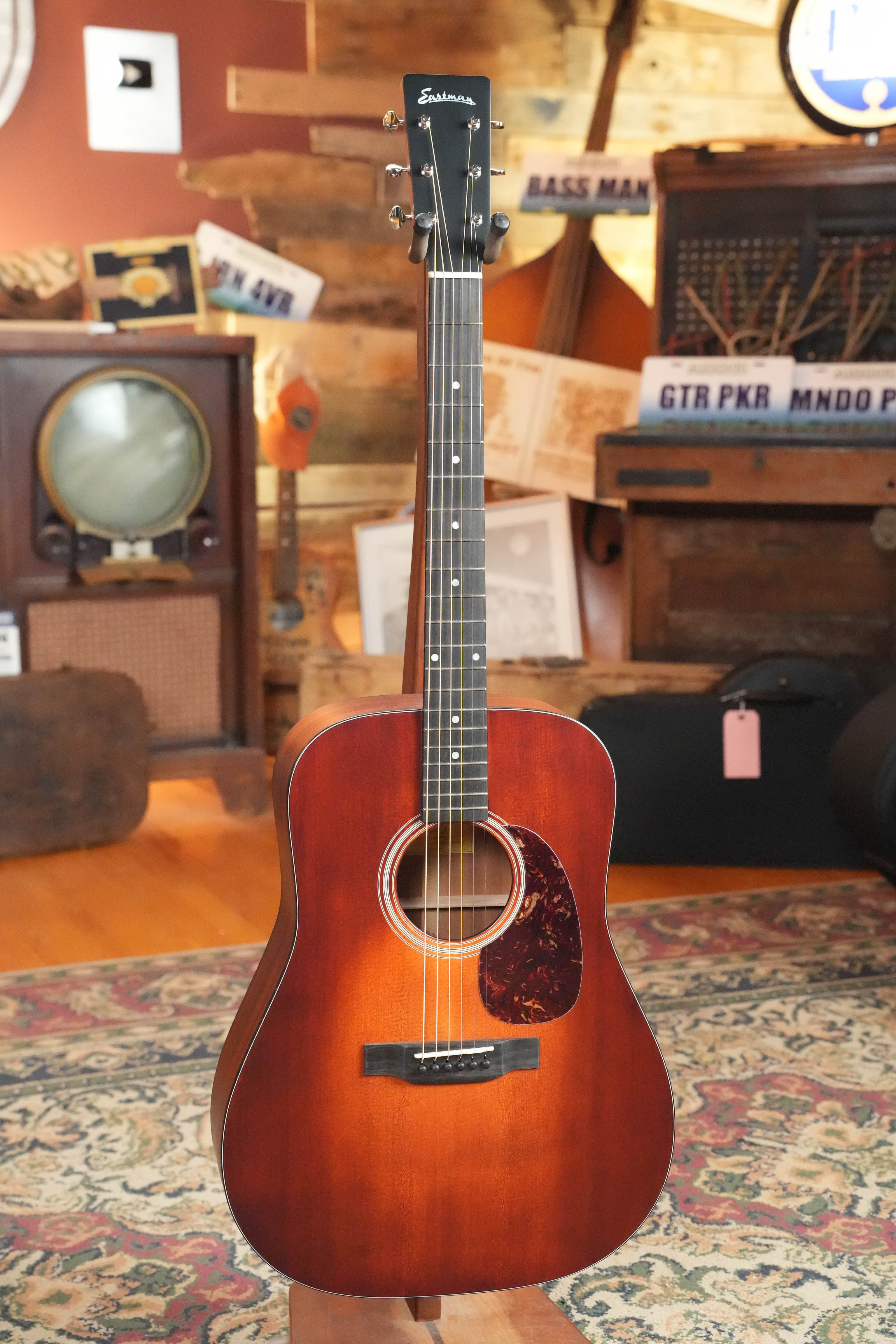 An Eastman E1D-CLA Classic Finish Dreadnought Acoustic Guitar by Eastman Guitars with a tortoiseshell pickguard stands upright on a patterned rug in a cozy, vintage-style room filled with musical instruments and retro decor.