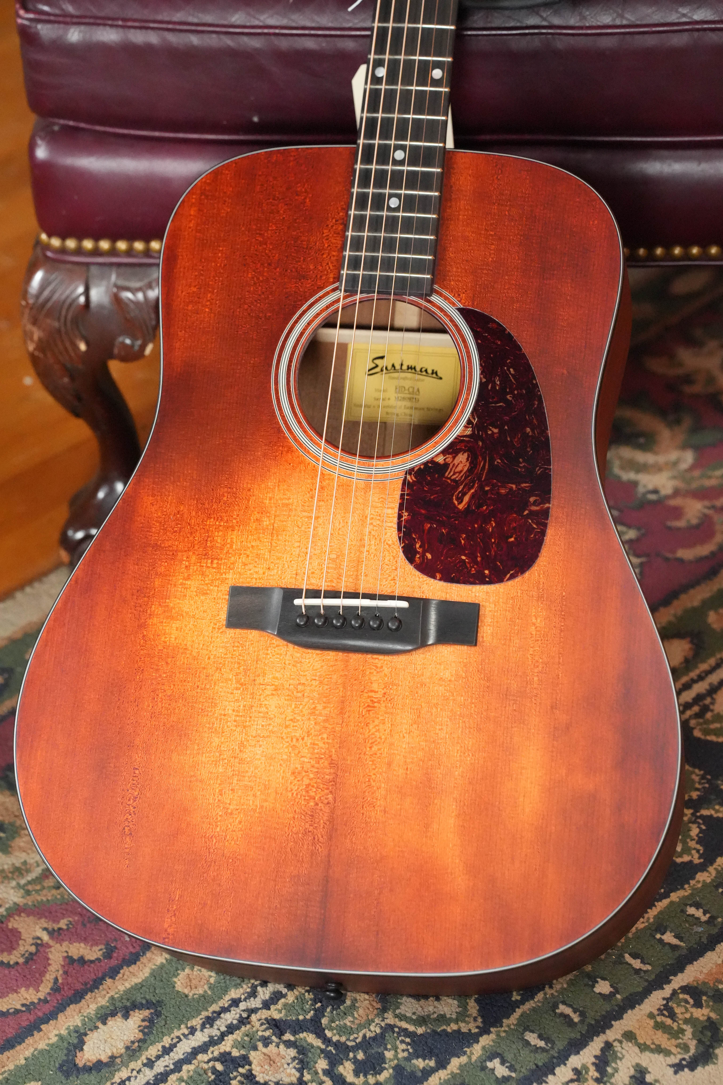 An Eastman E1D-CLA Classic Finish Dreadnought Acoustic Guitar #9753 from Eastman Guitars, with a solid Sitka spruce top and hand-scalloped X-bracing, rests on a patterned rug in front of a dark red tufted leather chair.