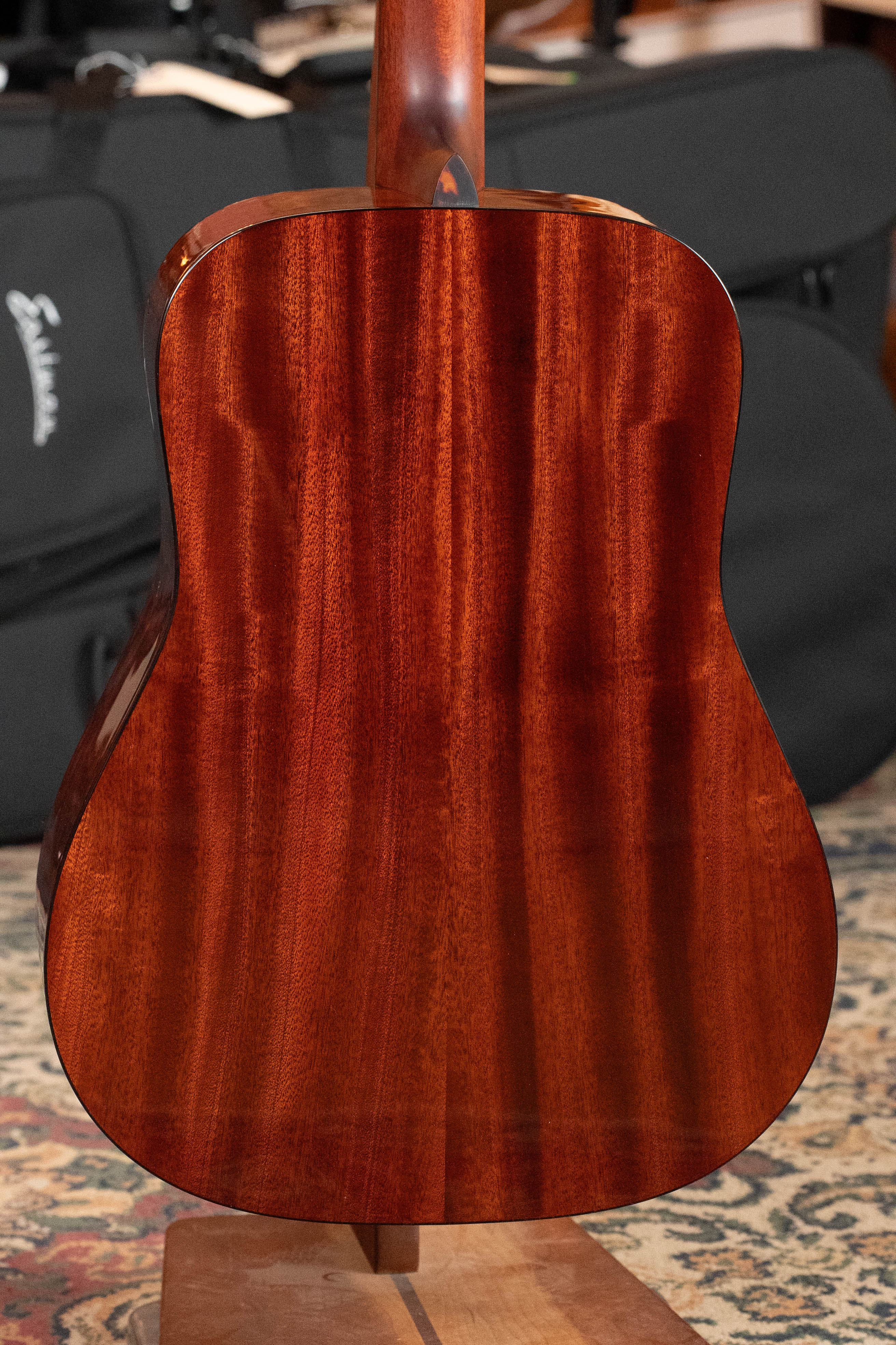 The image shows the back of an Eastman Guitars E1D-DLX Sitka/Sapele Dreadnought Guitar w/Fishman Pickup (#5656) with a polished reddish-brown finish, resting on a stand. A black case is in the background on a patterned rug.