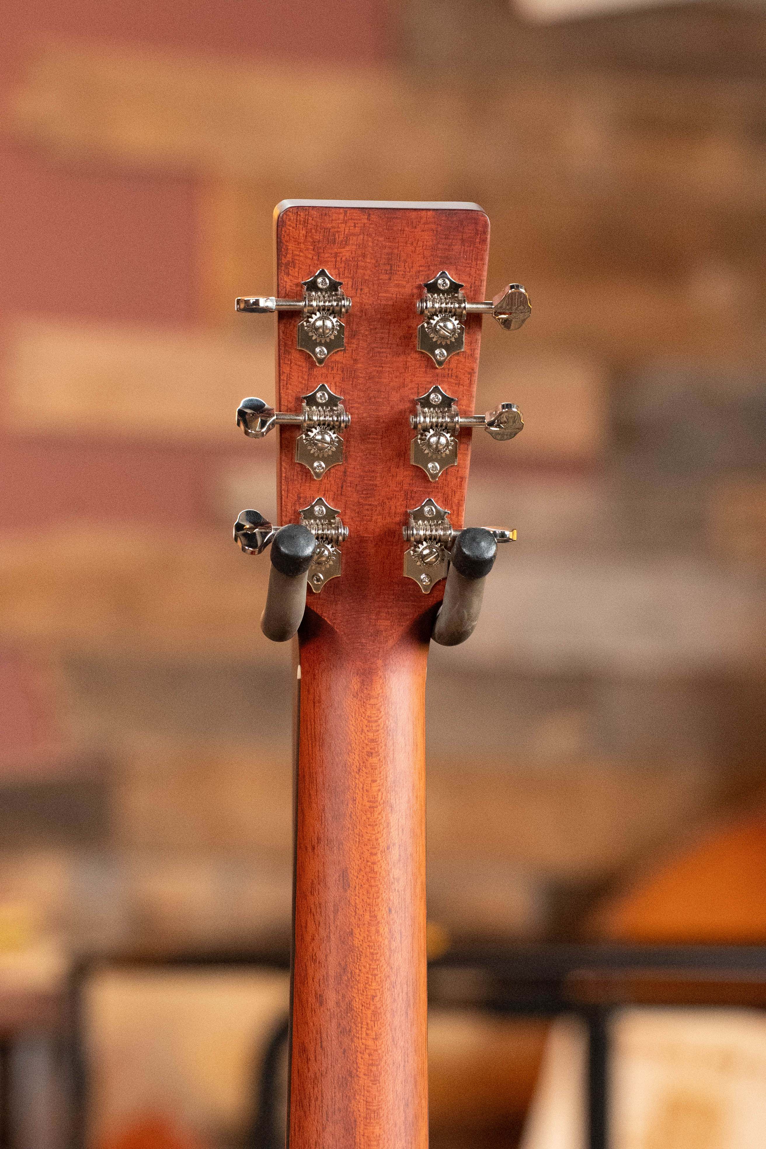 Close-up of the back of an Eastman E1D-DLX Sitka/Sapele Dreadnought Guitar by Eastman Guitars, showing six metal tuning pegs on a black hanger, with a warm wood and brick background.