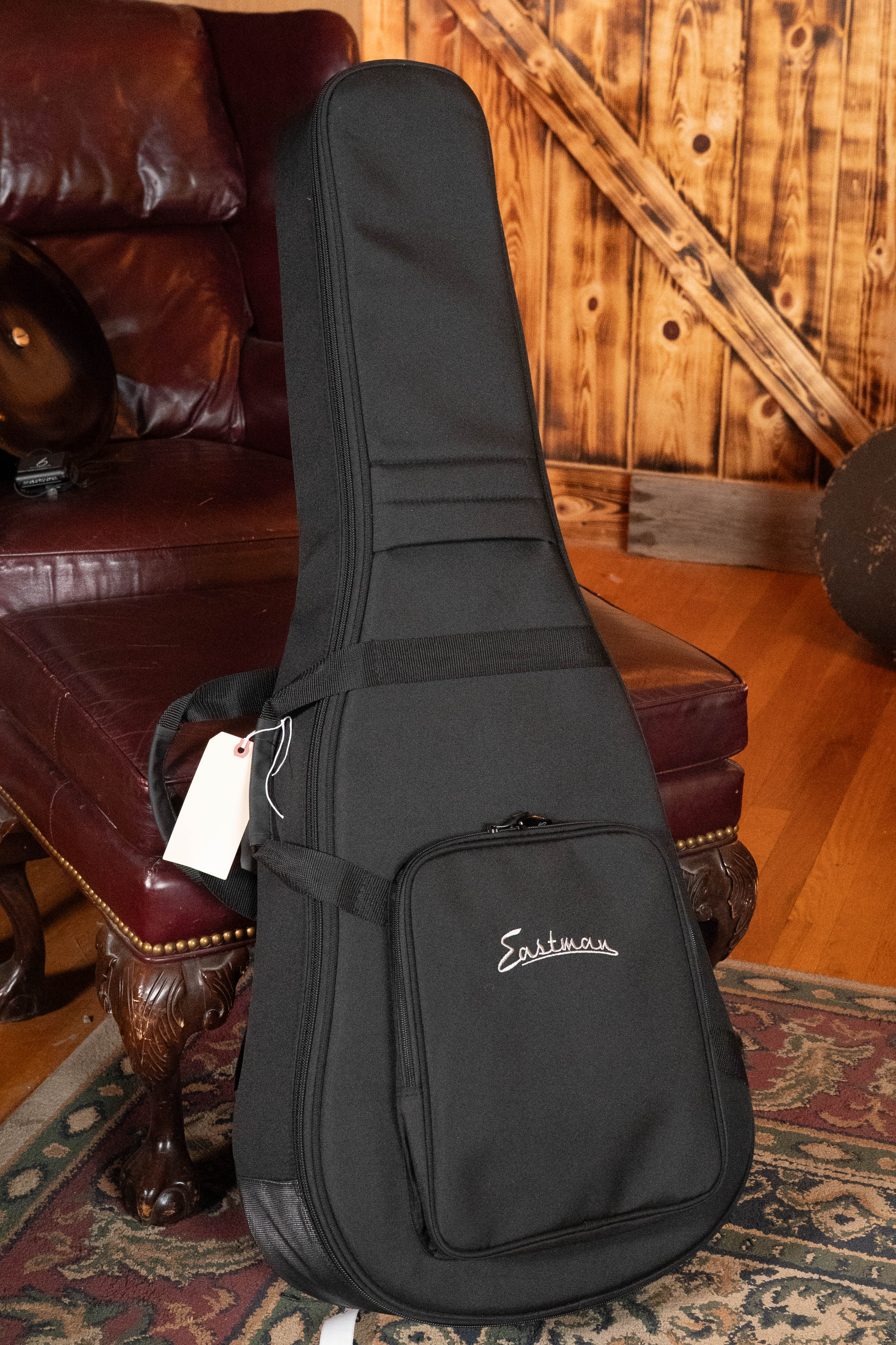 A black Eastman Guitars gig bag, made for the Eastman E1D-DLX Sitka/Sapele Dreadnought Guitar w/Fishman Pickup #5656, stands upright by a burgundy leather armchair in a cozy wood-paneled room, with a white tag on its handle.