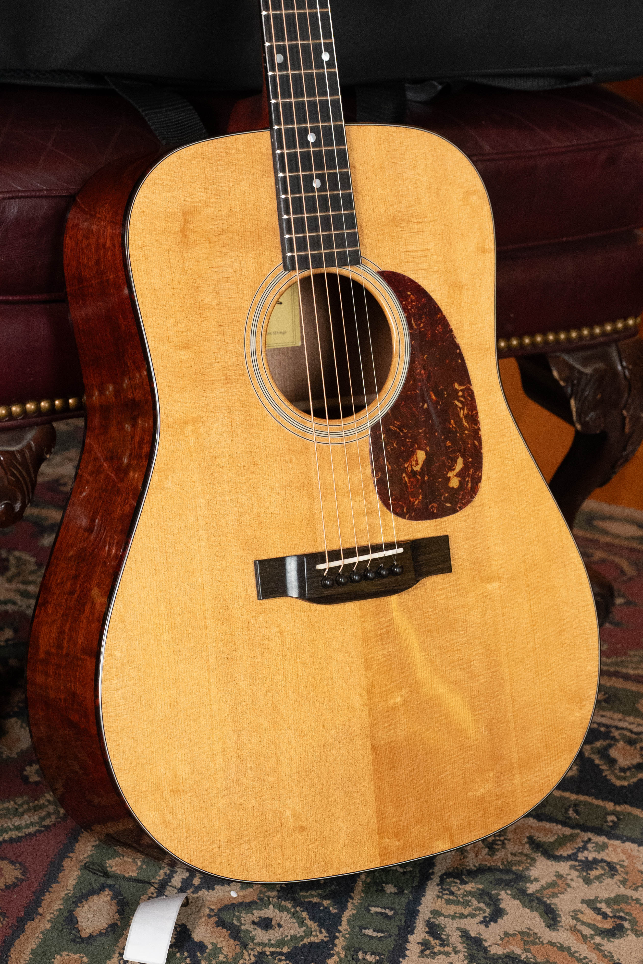 A close-up of the Eastman Guitars E1D-DLX Sitka/Sapele Dreadnought Guitar w/Fishman Pickup #5656, featuring a light wood top and dark sides, resting on an ornate rug before a maroon leather chair with brass studs.