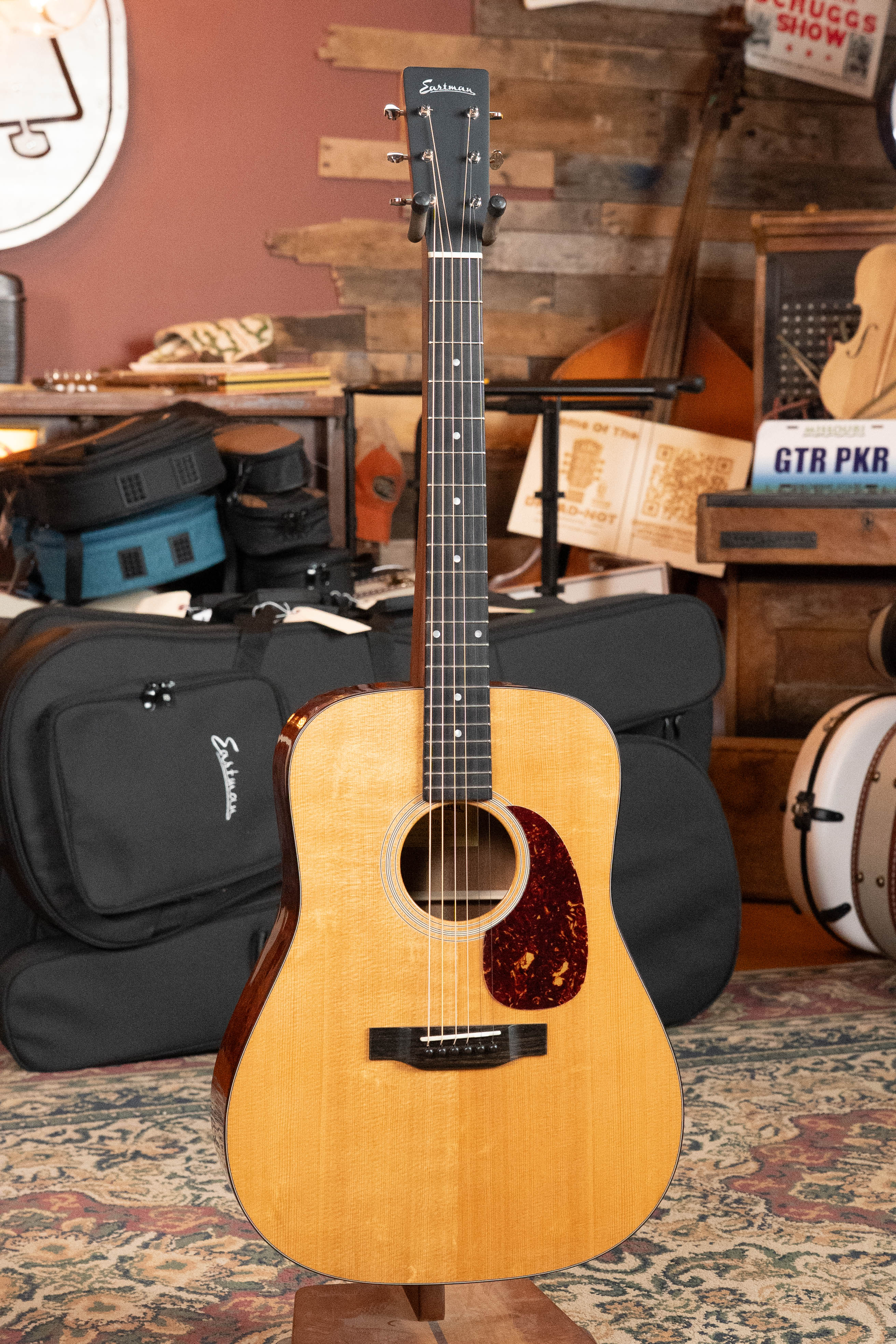An Eastman Guitars E1D-DLX Sitka/Sapele Dreadnought w/Fishman Pickup #5656 stands upright in a cozy, rustic room with musical instruments, gear bags, wooden shelves, and a carpeted floor.