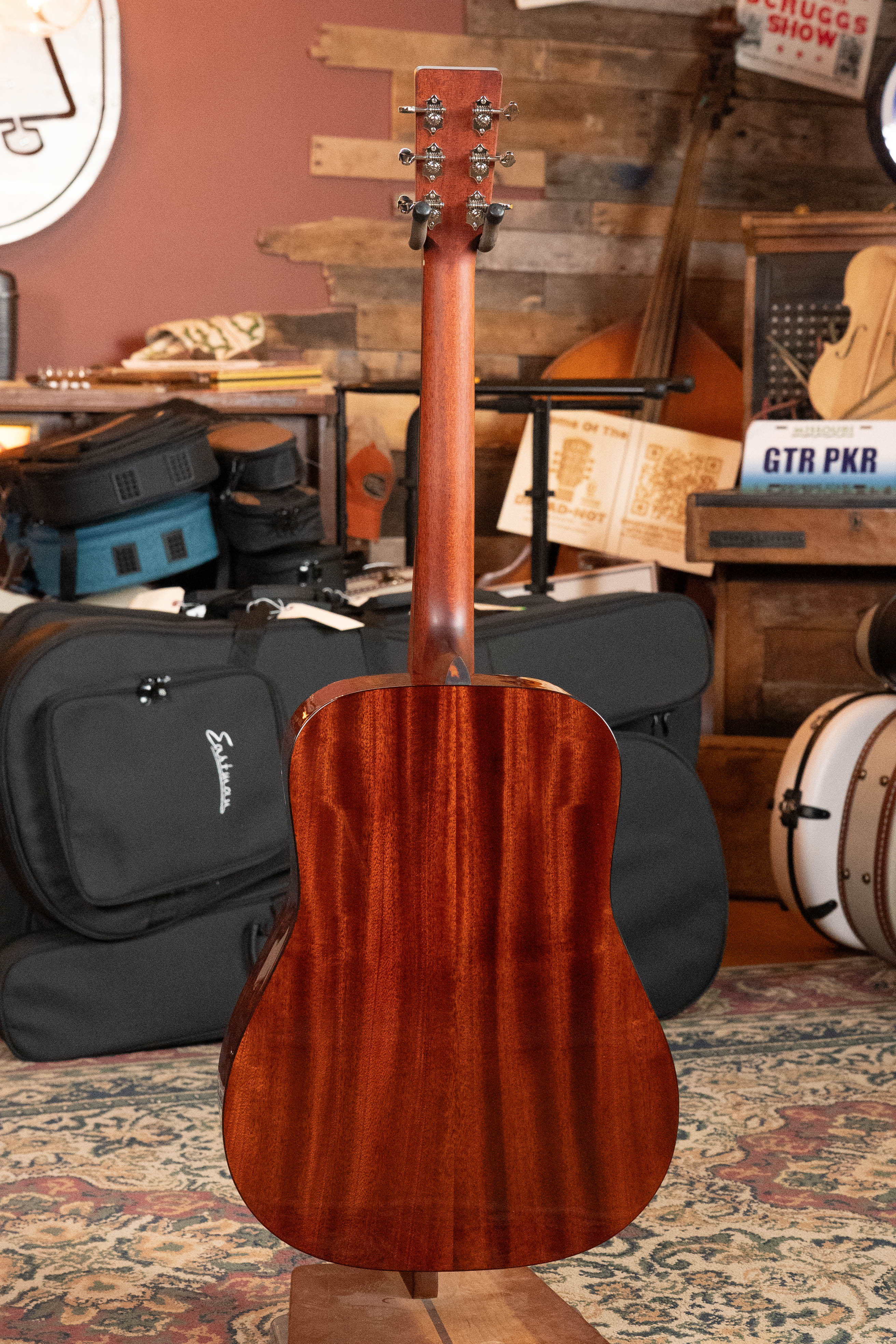 An Eastman Guitars E1D-DLX Sitka/Sapele Dreadnought Guitar w/Fishman Pickup #5656, featuring a polished brown finish, stands upright in a cozy room with musical instruments and wooden shelves in the background.