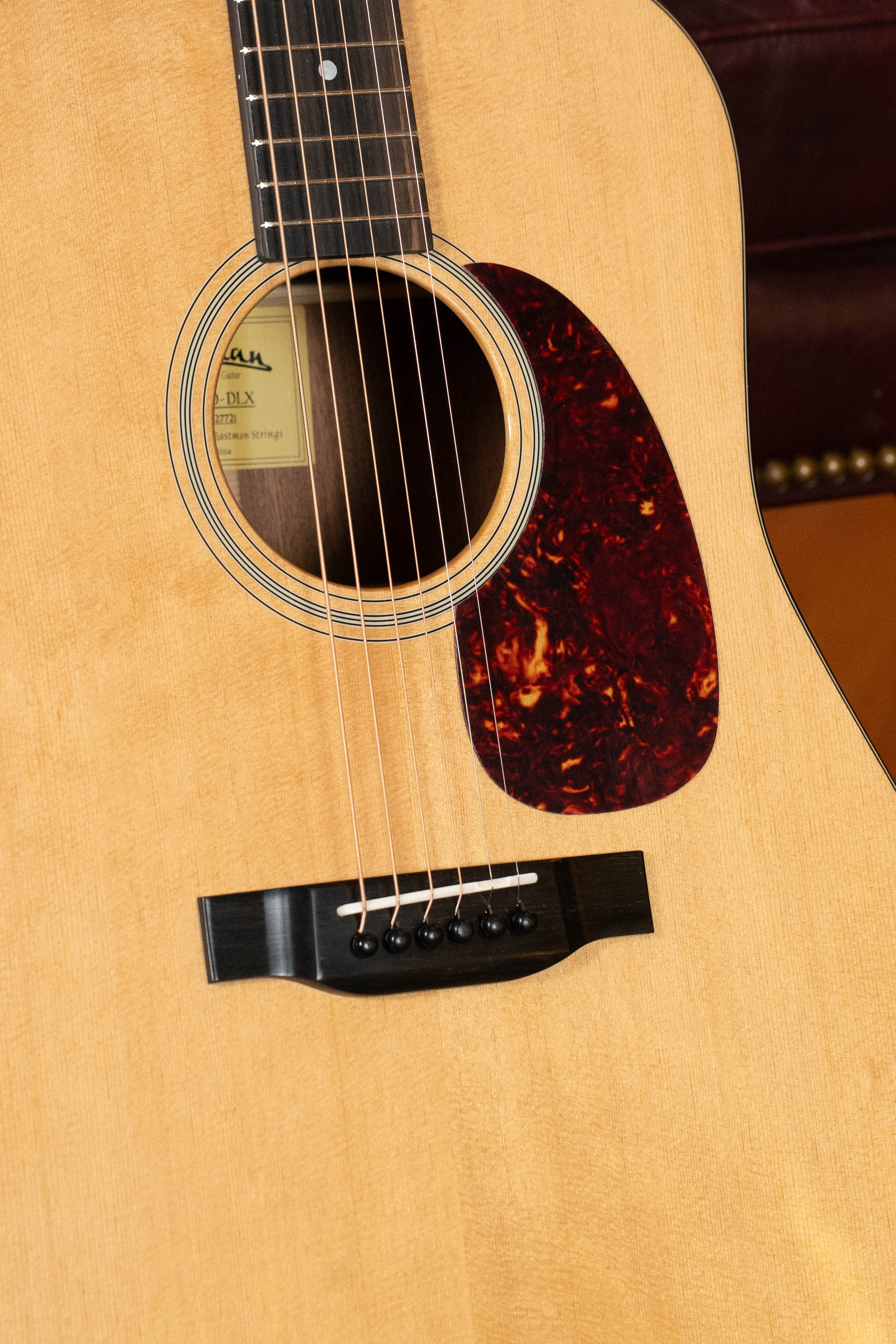A close-up of the Eastman Guitars E1D-DLX Sitka/Sapele Dreadnought Guitar w/Fishman Pickup #7721, highlighting its solid Sitka spruce top, strings, sound hole, pickguard, and fretboard in front of a brown leather chair.