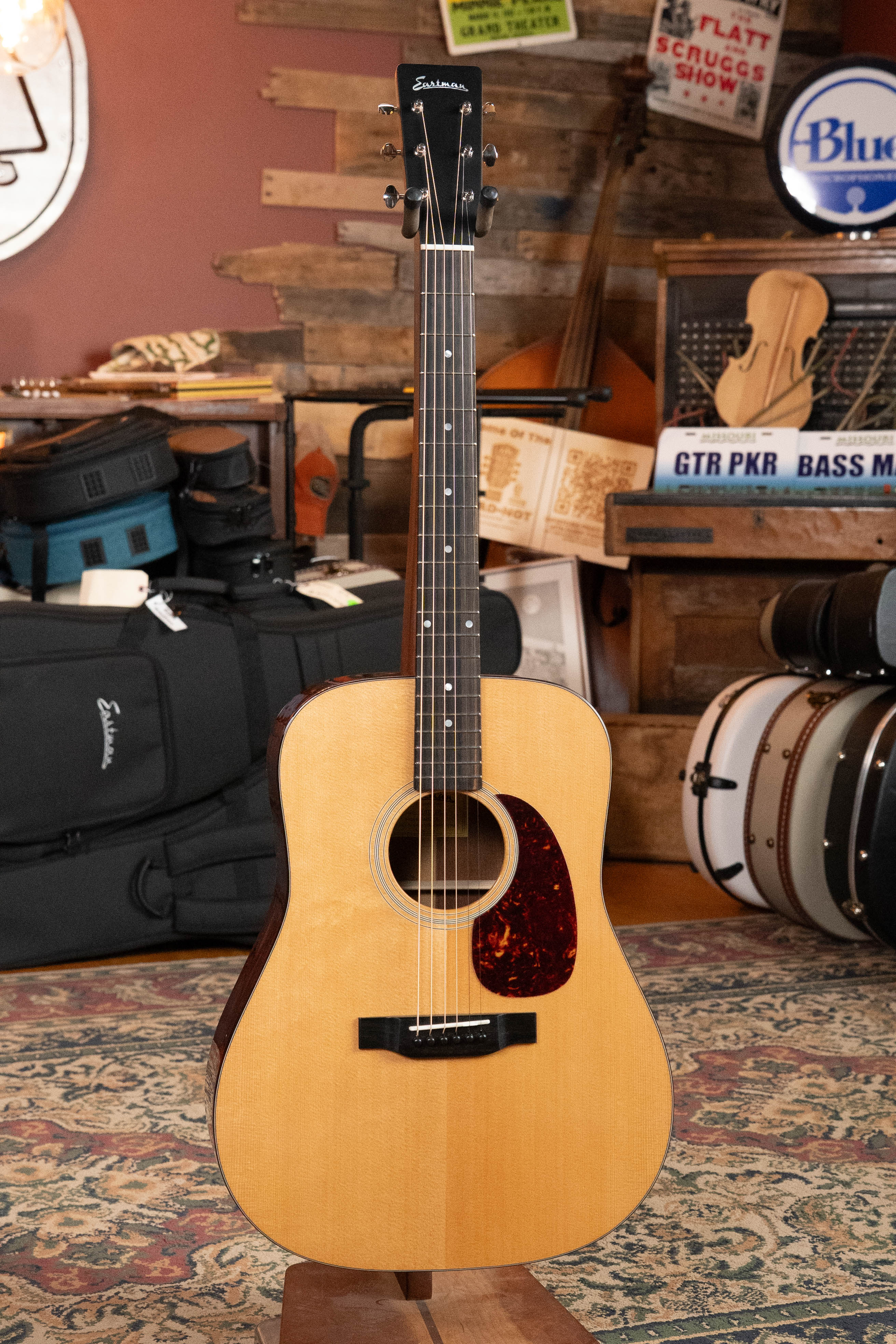 An Eastman Guitars E1D-DLX Sitka/Sapele Dreadnought Guitar w/Fishman Pickup #7721 stands upright on a stand in a cozy room filled with musical instruments, cases, and vintage signs in the background.