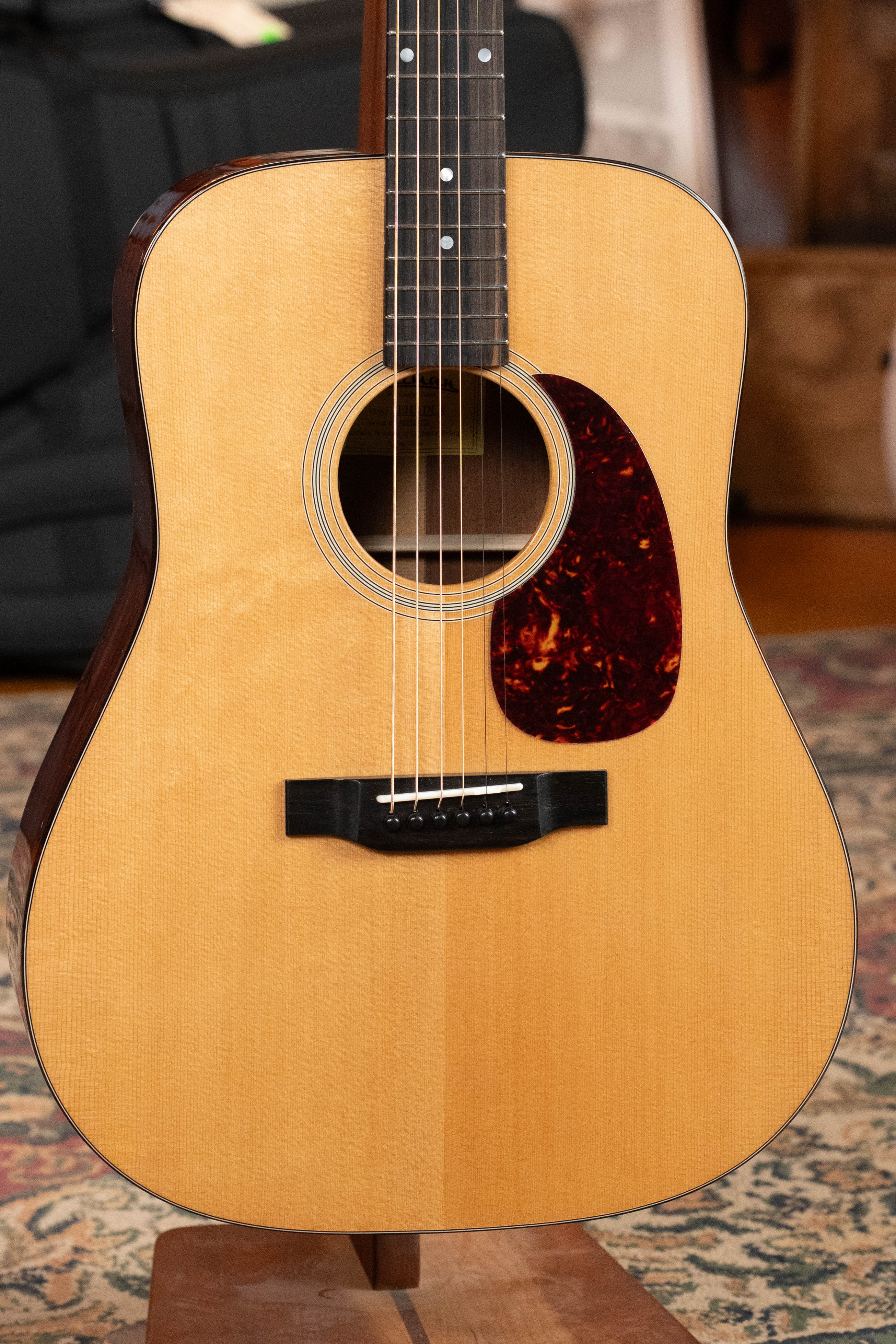 A close-up of an Eastman Guitars E1D-DLX Sitka/Sapele Dreadnought Guitar w/Fishman Pickup #7721, with solid Sitka spruce top and dark pickguard, standing on a wooden stand. Patterned carpet and blurred furniture are in the background.