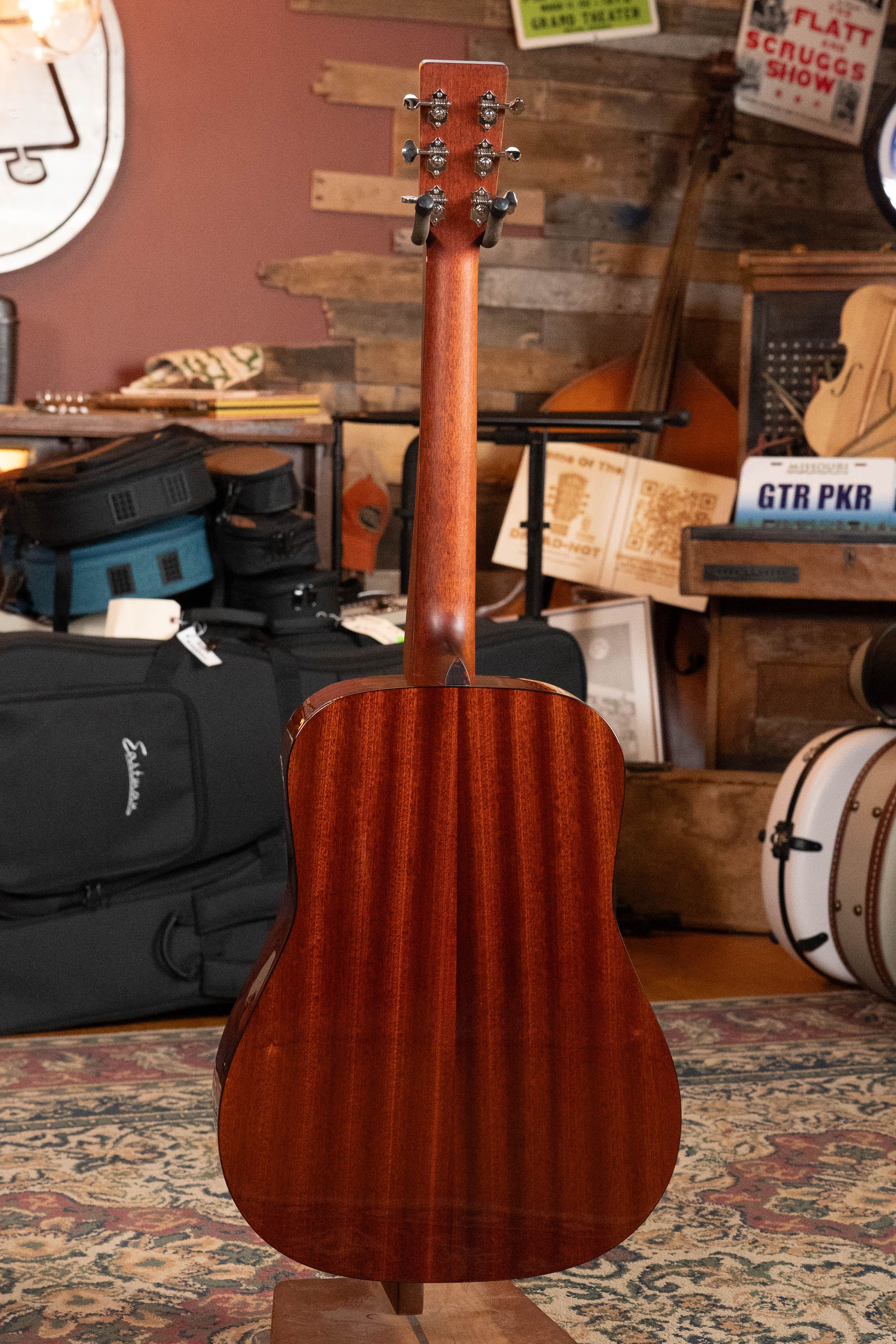 An Eastman Guitars E1D-DLX Sitka/Sapele Dreadnought Guitar w/Fishman Pickup (#7721) stands upright, back facing out, in a cozy room filled with musical instruments, equipment cases, books, and posters.