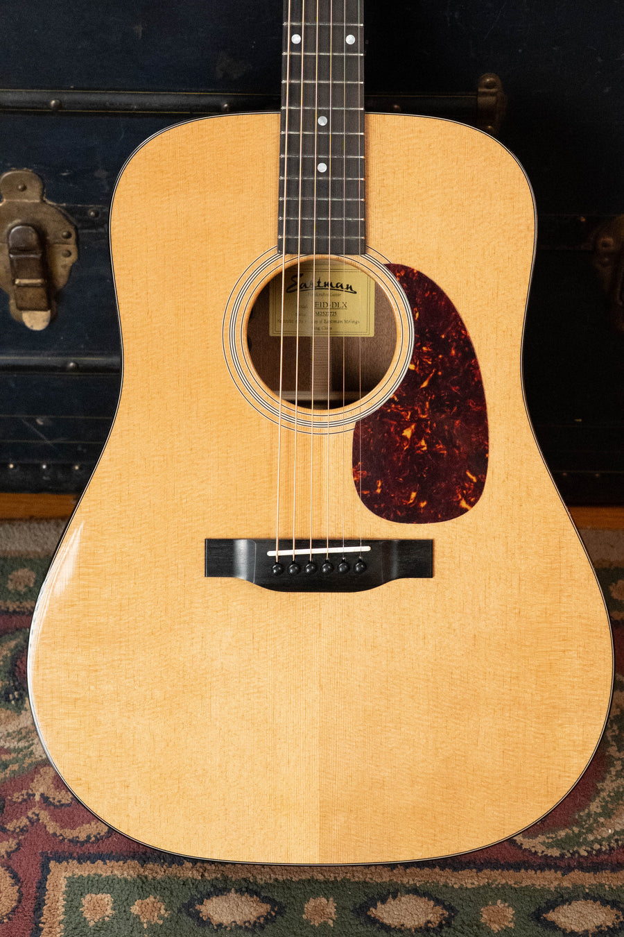 A close-up of the Eastman E1D-DLX Sitka/Sapele Dreadnought Guitar by Eastman Guitars, featuring a natural finish, dark pickguard, and Fishman pickup (#7725), standing upright on a patterned rug with a dark trunk behind it.