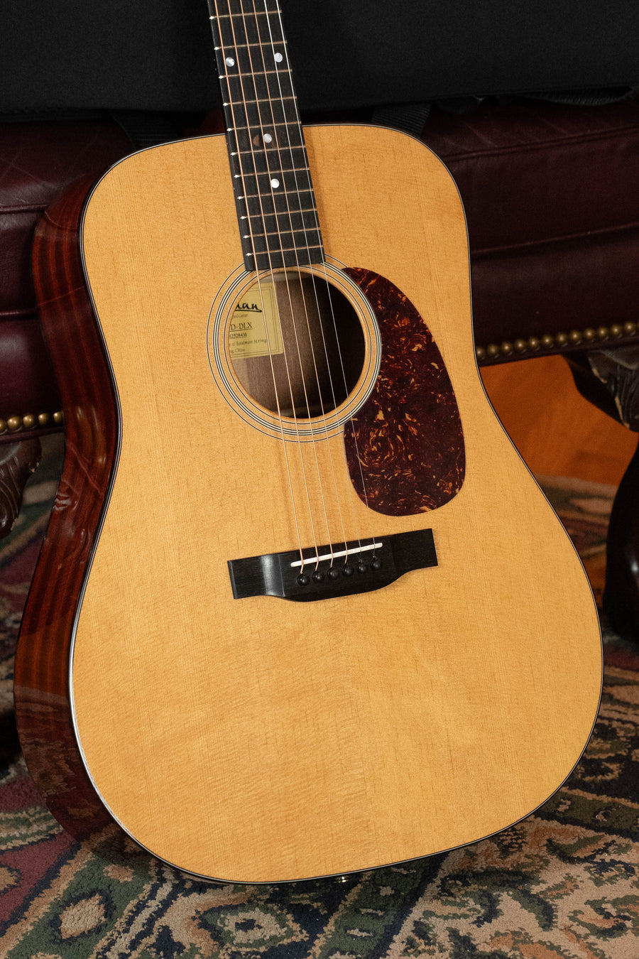 A close-up of the Eastman Guitars E1D-DLX Sitka/Sapele Dreadnought Guitar w/Fishman Pickup #8436, featuring a light top, dark sapele sides, and tortoiseshell pickguard, resting on a patterned rug in front of a leather couch.