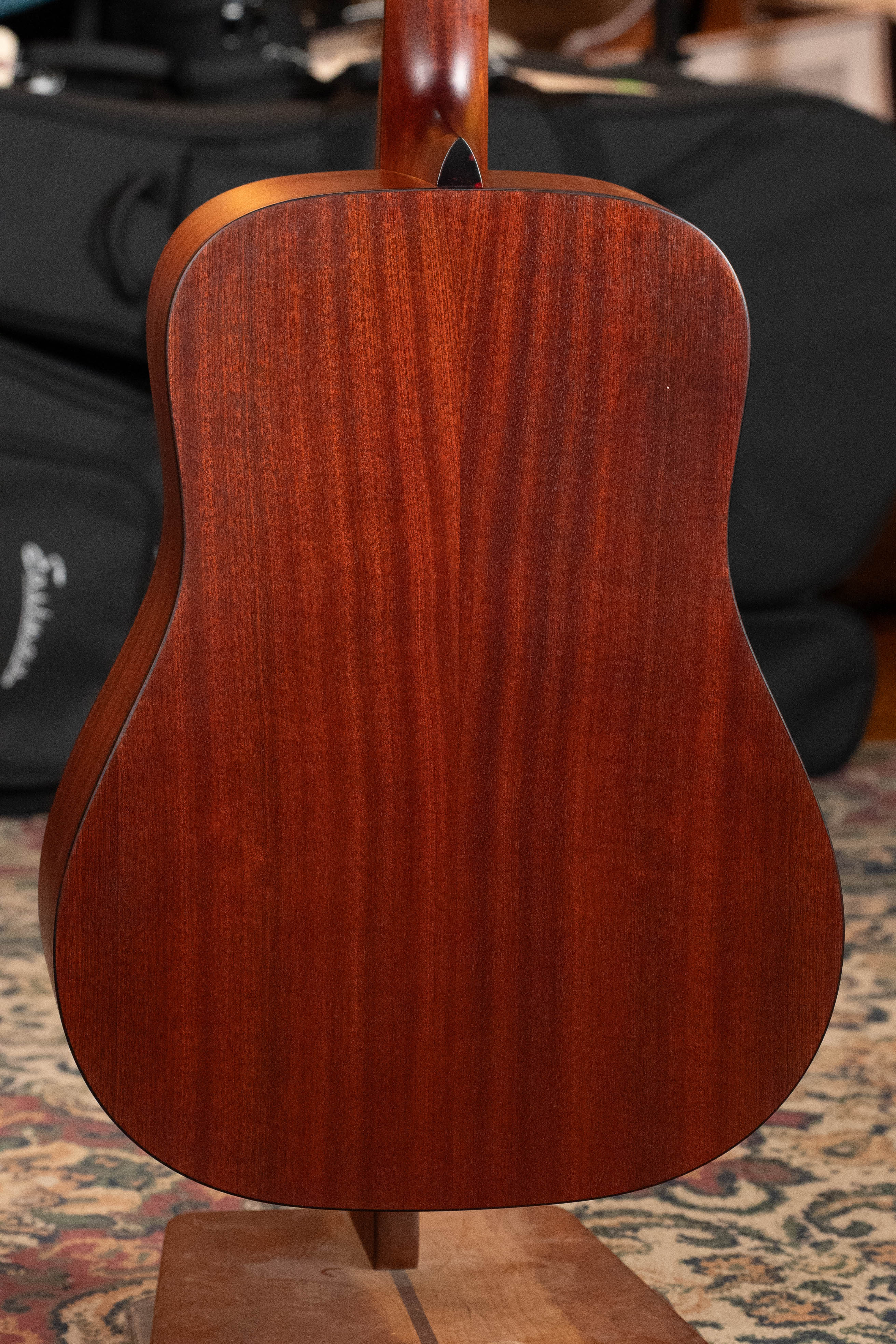 The back of an Eastman Guitars E1D Sitka/Sapele Dreadnought Acoustic Guitar #6855 is shown upright on a stand, highlighting its rich reddish-brown finish, with a patterned rug and a black guitar case in the background.