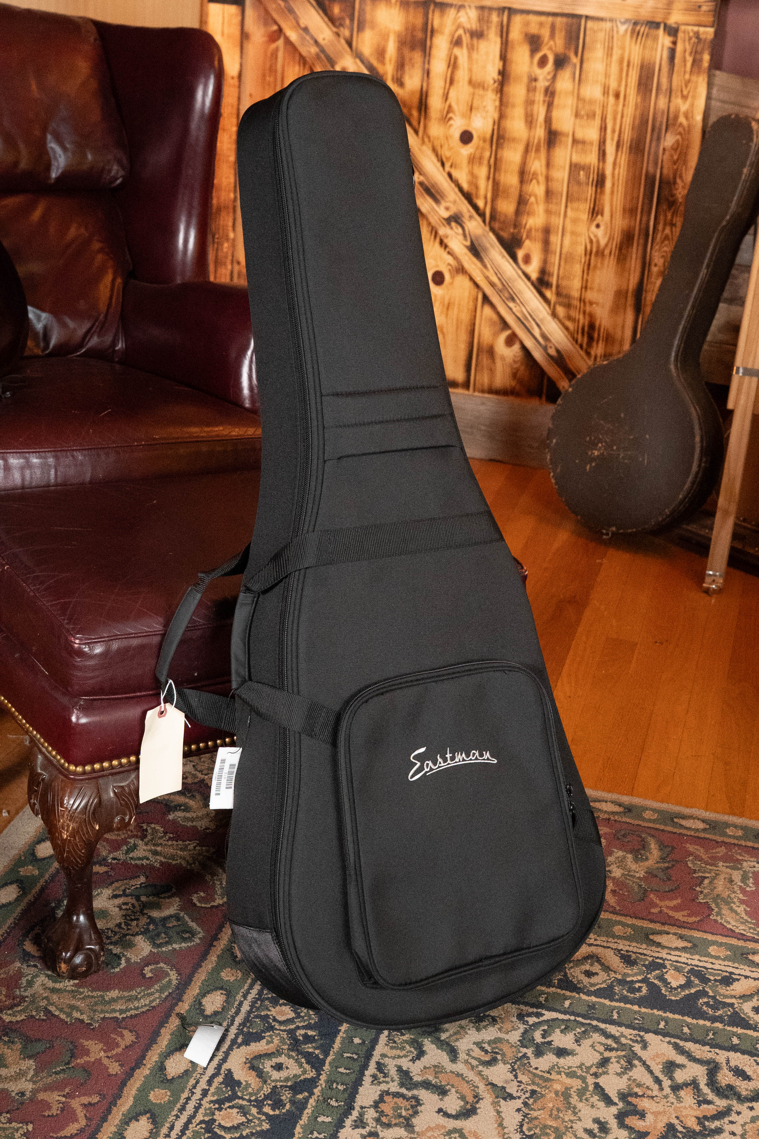 A black Eastman Guitars case, ideal for the Eastman E1D Sitka/Sapele Dreadnought Acoustic Guitar #6855, stands upright on a patterned rug in a cozy, wood-paneled room with a leather couch and another acoustic guitar case nearby.
