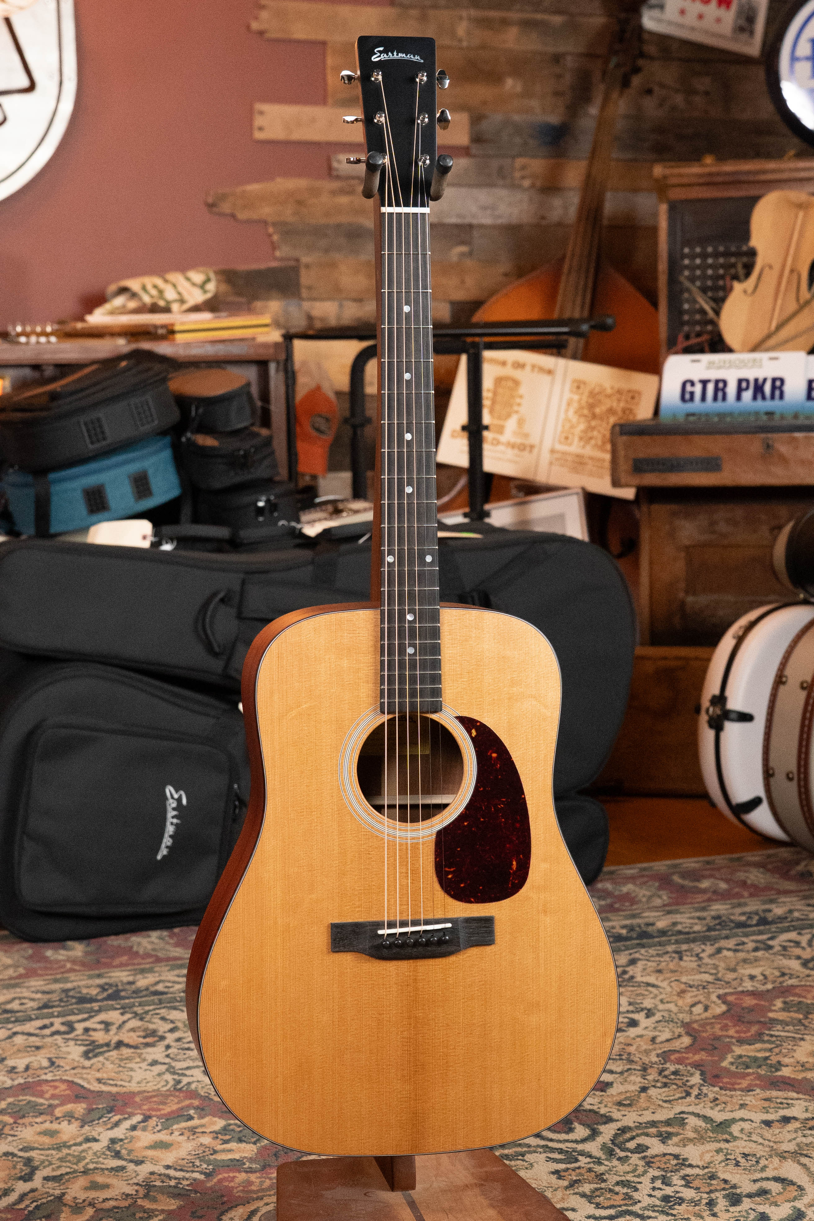 An Eastman Guitars E1D Sitka/Sapele Dreadnought Acoustic Guitar (#6855) with a natural wood finish and tortoiseshell pickguard stands upright in a cozy, rustic room filled with musical instruments and cases.
