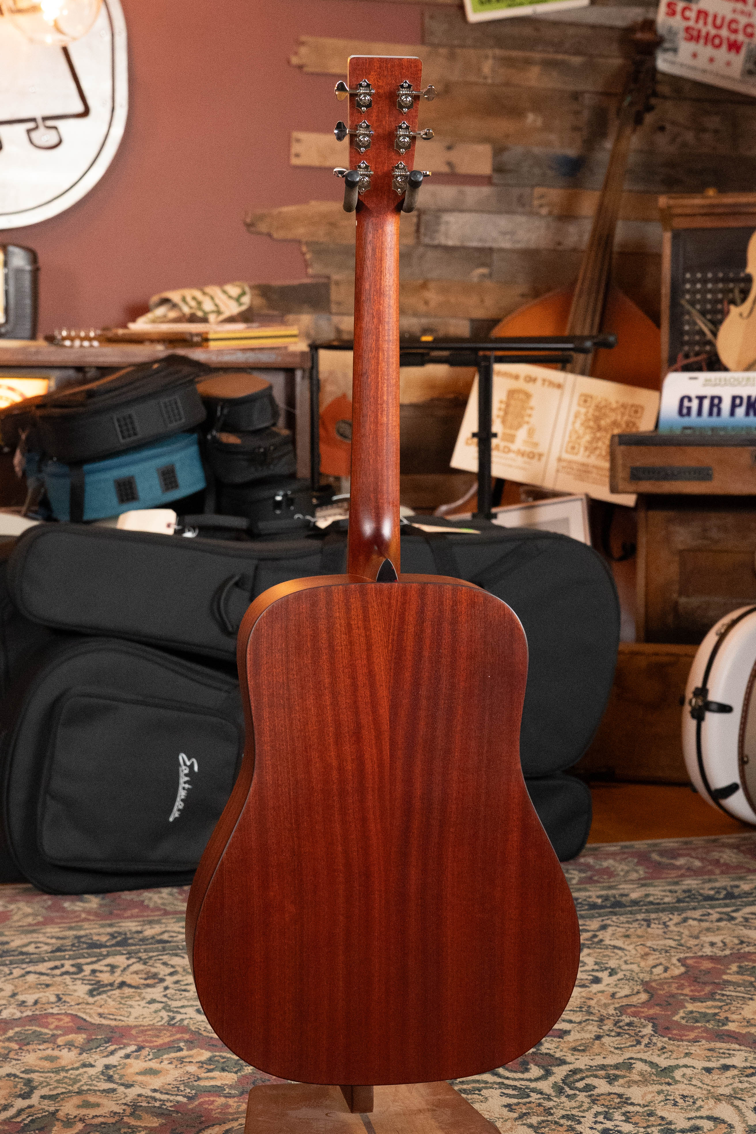 An Eastman Guitars E1D Sitka/Sapele Dreadnought Acoustic Guitar (#6855) with a smooth, reddish-brown finish stands upright in a room filled with instrument cases, books, and other stringed instruments.
