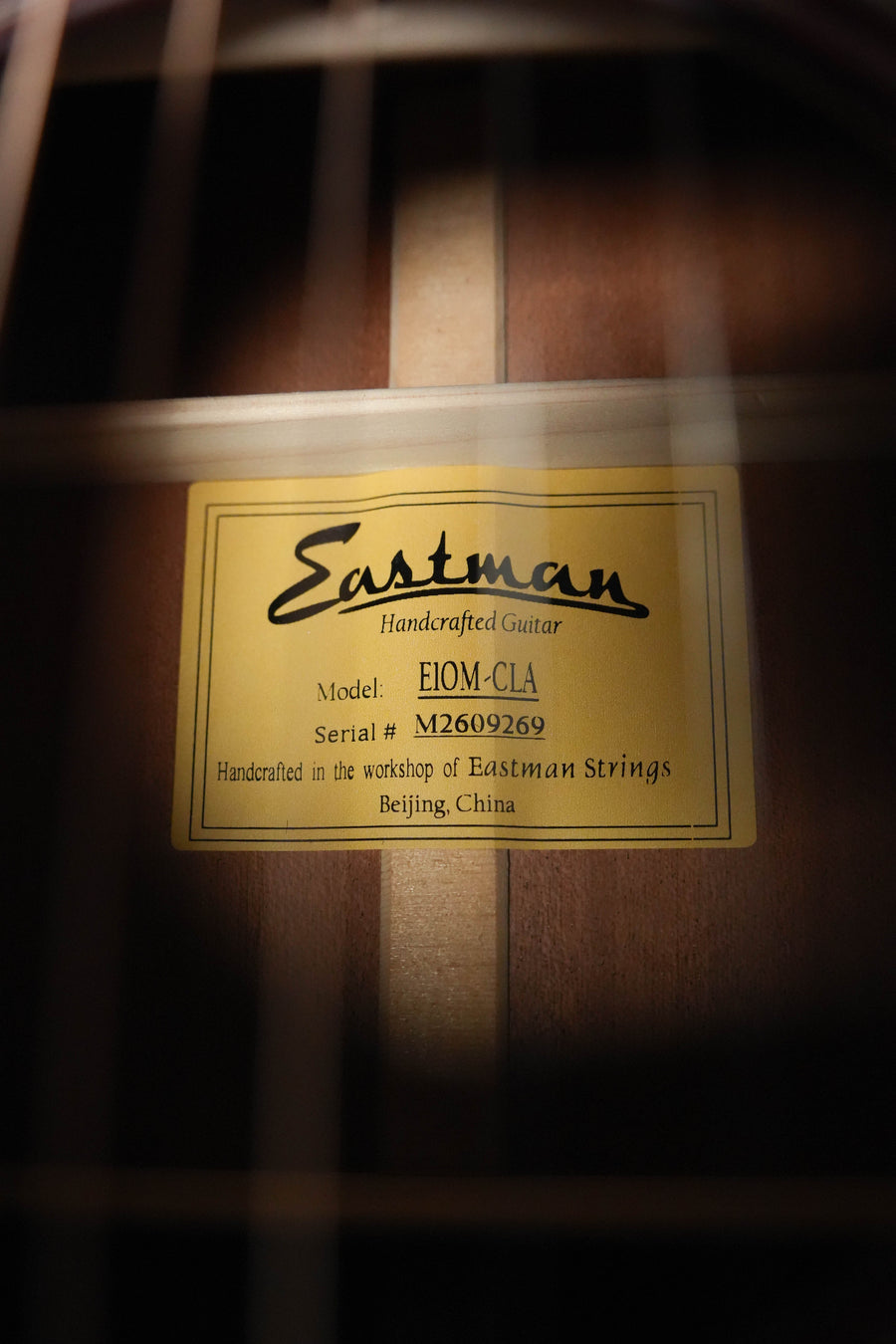 Close-up of a yellow label inside an Eastman Guitars E1OM-CLA All Solid Sitka/Sapele Orchestra Model Acoustic Guitar #9269, handcrafted in Beijing, China. The solid Sitka spruce top and guitar strings are visible in the foreground.