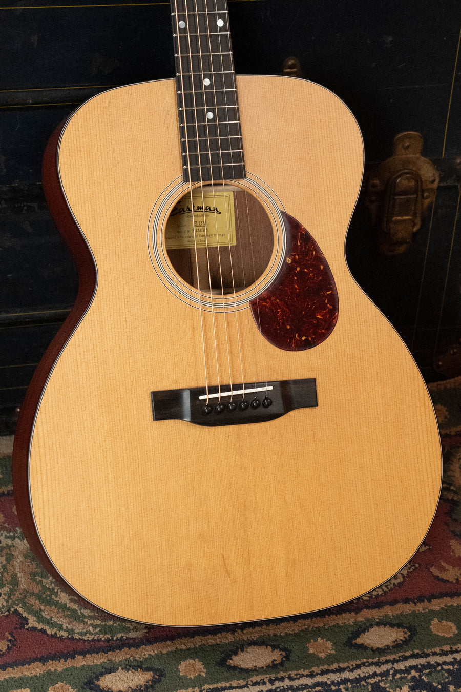 A close-up of an Eastman Guitars E1OM All Solid Sitka/Sapele Acoustic Guitar #7765 featuring a Sitka spruce top, dark sapele sides, tortoiseshell pickguard, and round rosette, resting on a rug beside its dark brass-hardware case.