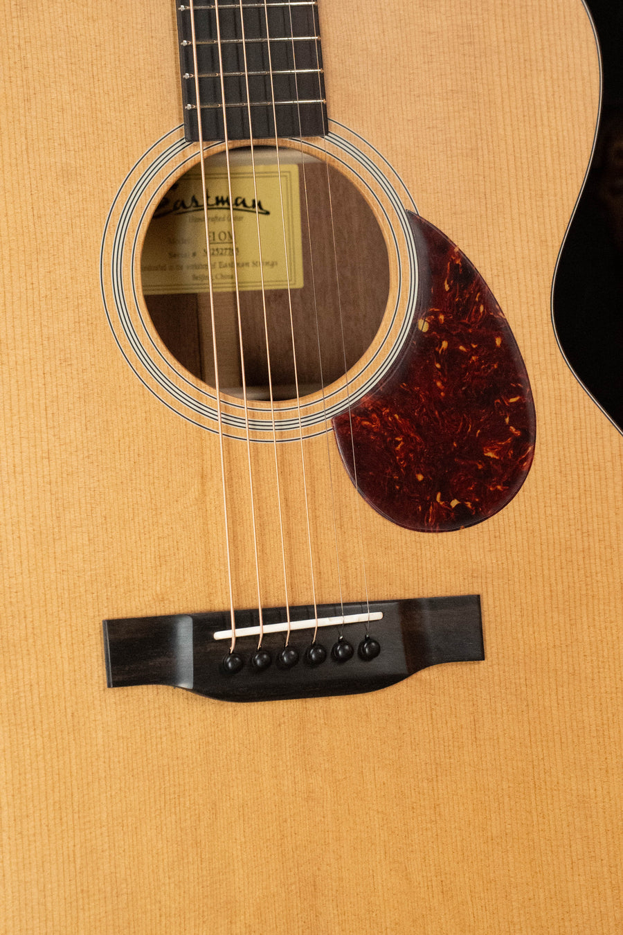 Close-up of the Eastman Guitars E1OM All Solid Sitka/Sapele Acoustic Guitar #7765 body, showing its solid Sitka spruce top, strings, soundhole label, black bridge with white pins, and reddish-brown pickguard on a light finish.