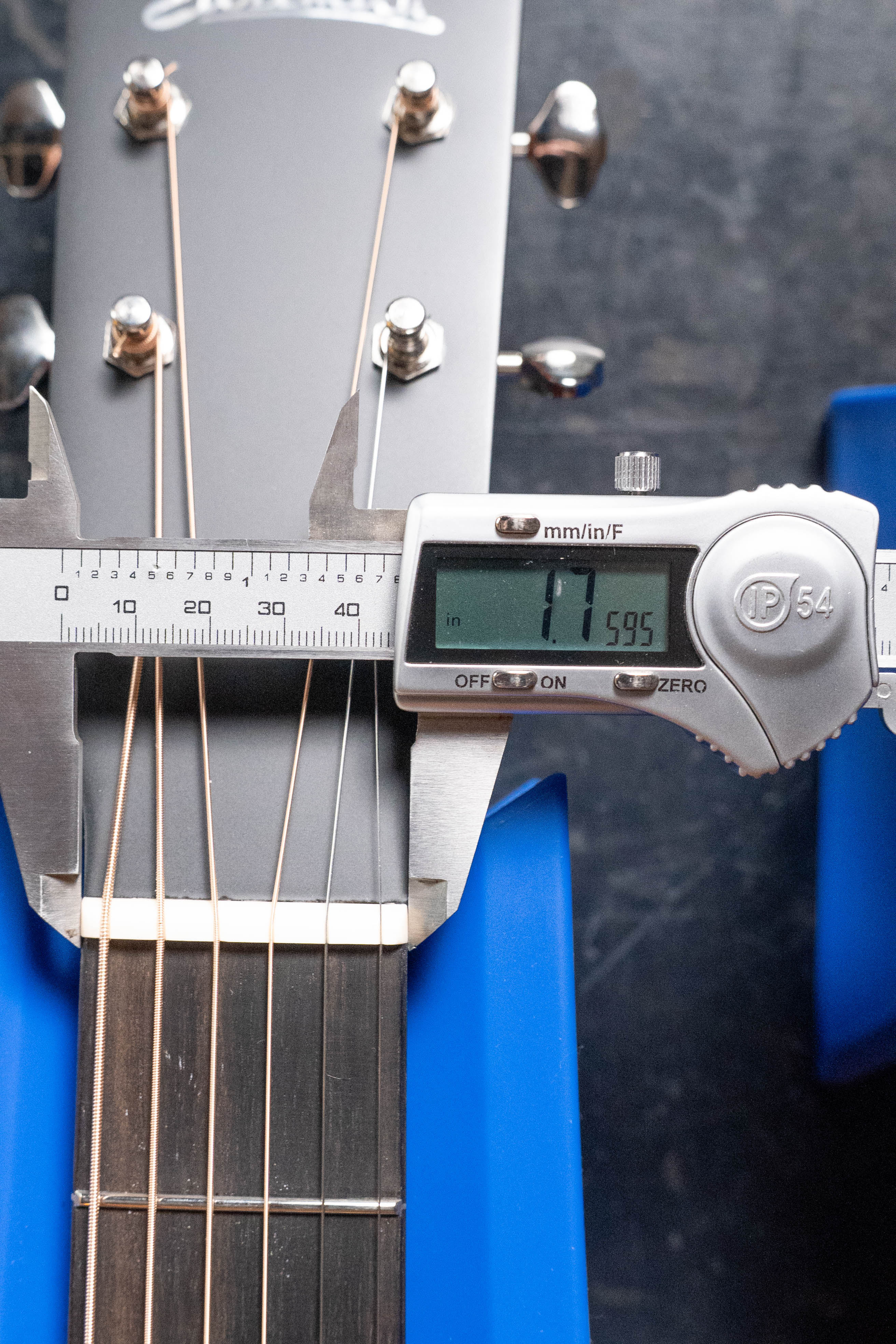 A digital caliper shows a 1.595-inch neck width near the headstock of an Eastman Guitars E1OM All Solid Sitka/Sapele Orchestra Model Acoustic Guitar #7102, which is topped with solid Sitka spruce and rests on a blue holder.