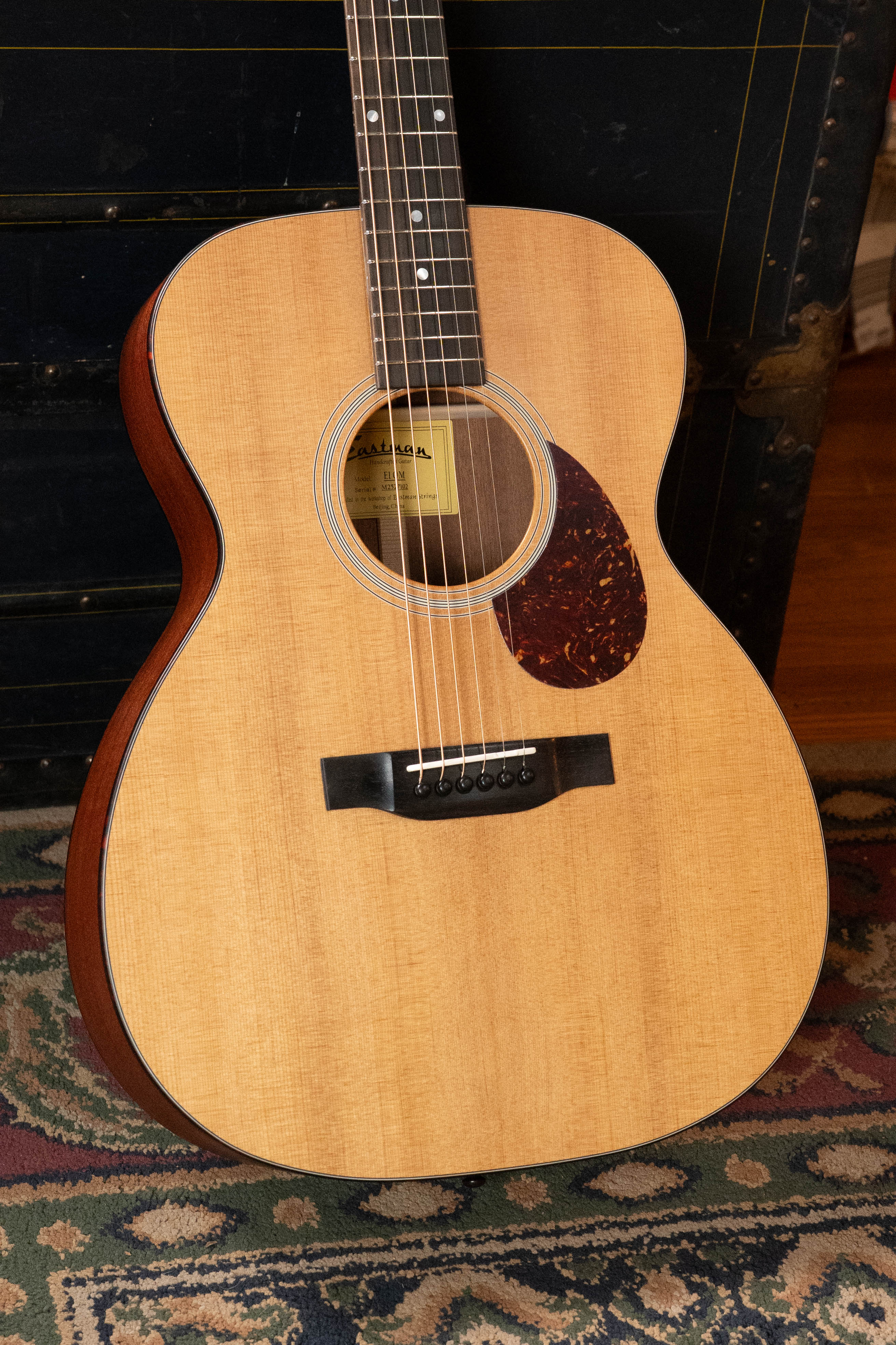 An Eastman Guitars E1OM All Solid Sitka/Sapele Orchestra Model Acoustic Guitar #7102 stands upright on a patterned rug, with a dark trunk and wooden floor in the background.