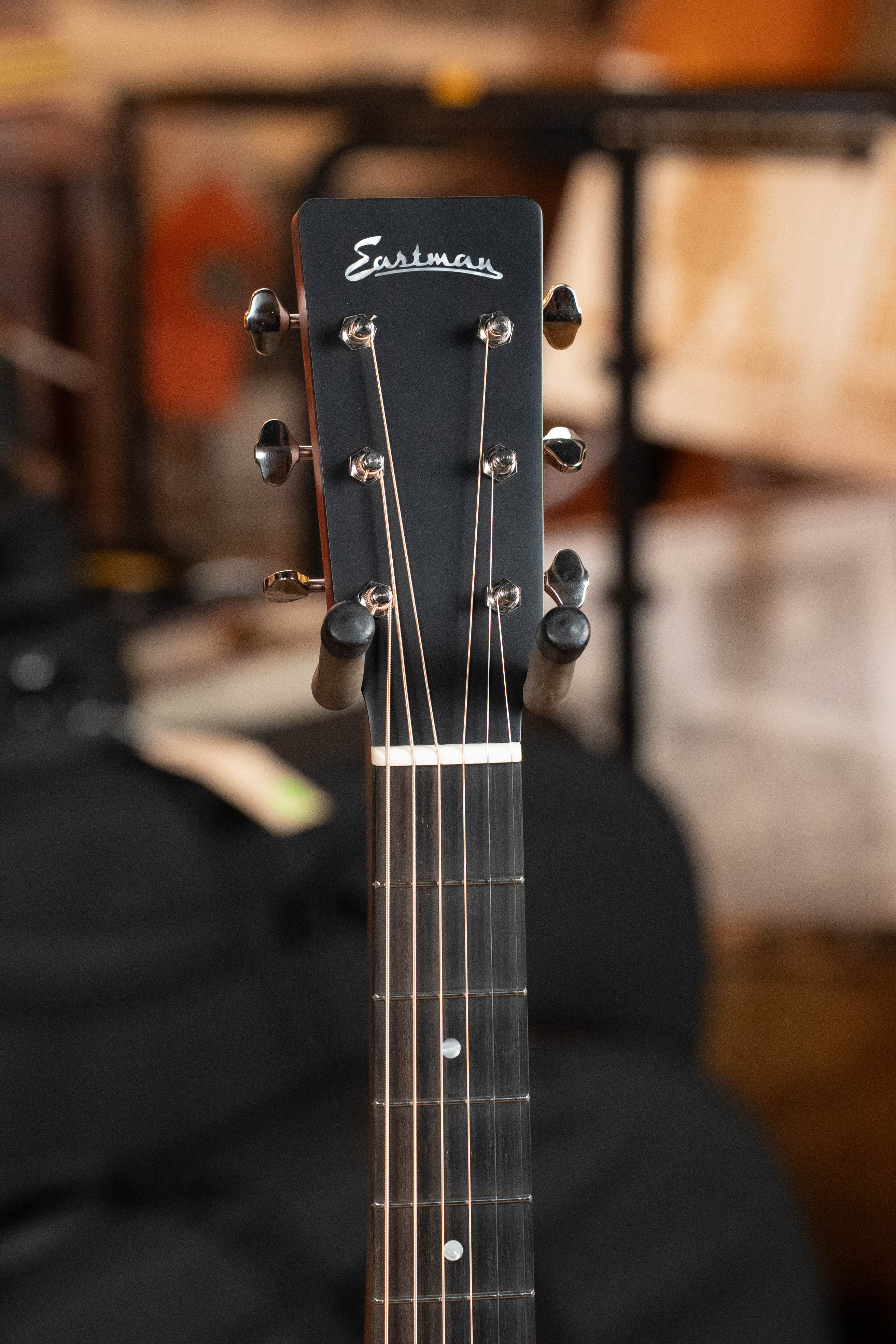 Close-up of the headstock on an Eastman Guitars E1OM All Solid Sitka/Sapele Orchestra Model Acoustic Guitar #7102, featuring tuning pegs and part of the fretboard. The blurred background shows indistinct objects and musical equipment.