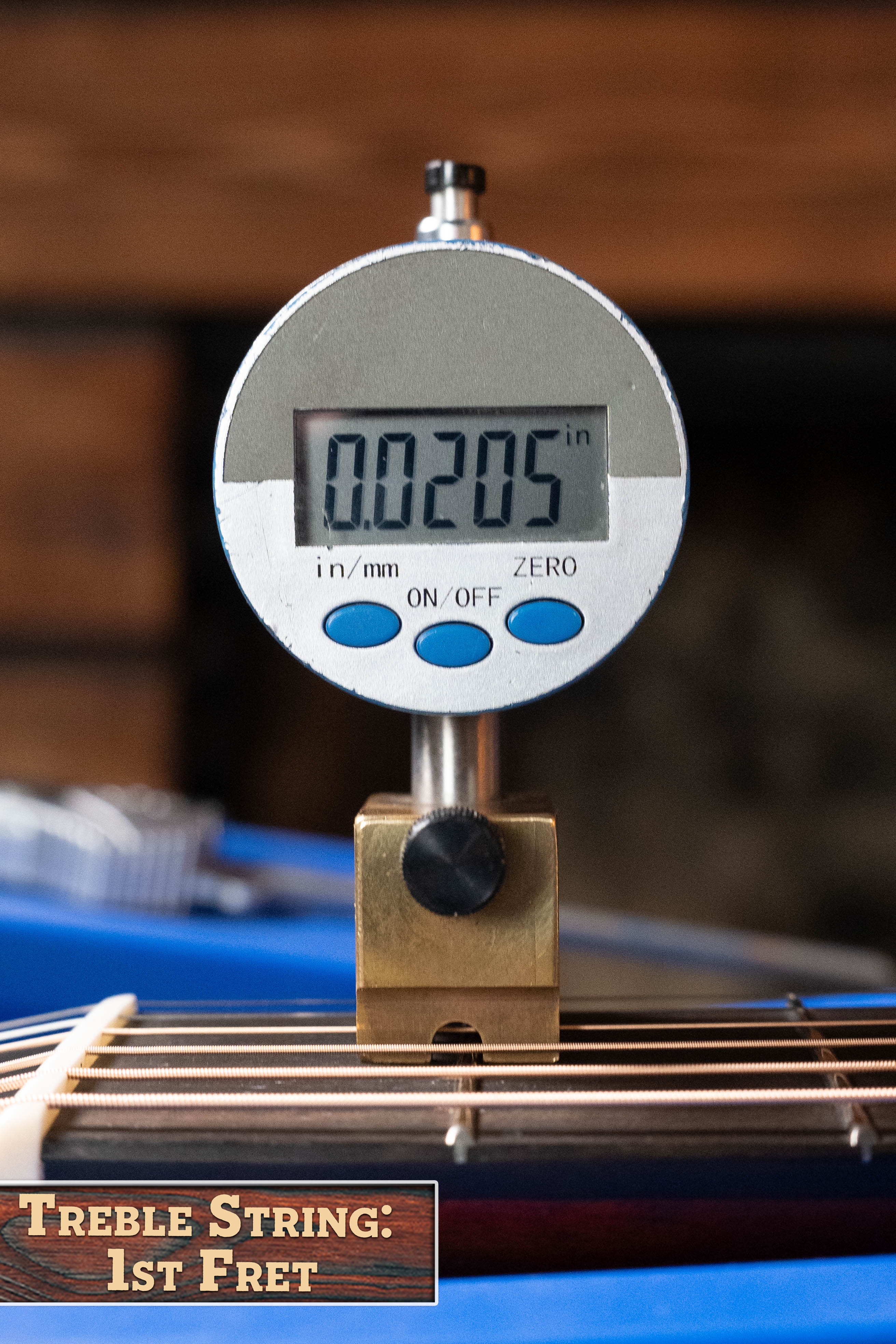A digital gauge shows 0.0205 inches above the first fret treble string on an Eastman Guitars E1OM All Solid Sitka/Sapele Orchestra Model Acoustic Guitar #7102, with a blurred wood background and "Treble String: 1st Fret" label visible.