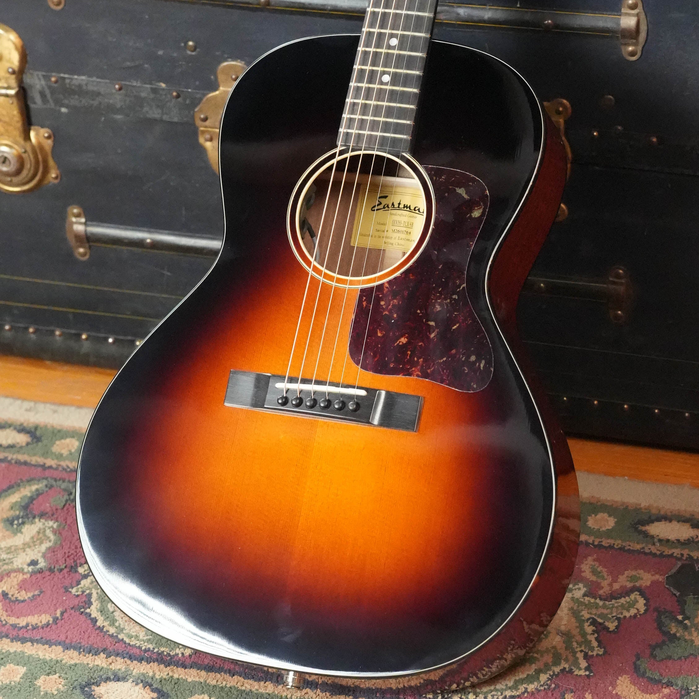 The Eastman Guitars E1OOSS-DLX-SB Sunburst 00 Acoustic-Electric Guitar #1764, featuring a mahogany neck and tortoiseshell pickguard, stands upright against a black trunk on a patterned rug.