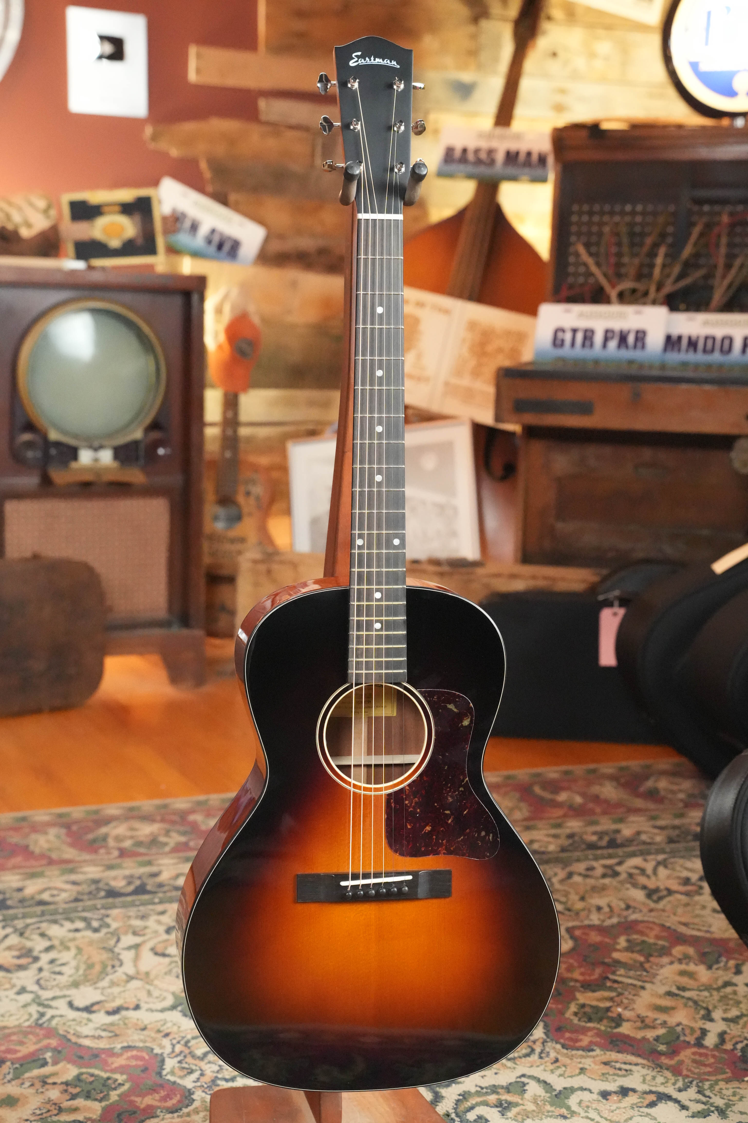 The Eastman Guitars E1OOSS-DLX-SB Sunburst 00 Acoustic-Electric Guitar #1764, featuring a mahogany neck, is displayed upright in a vintage room with an old TV, wooden furniture, rugs, and musical signs in the background.