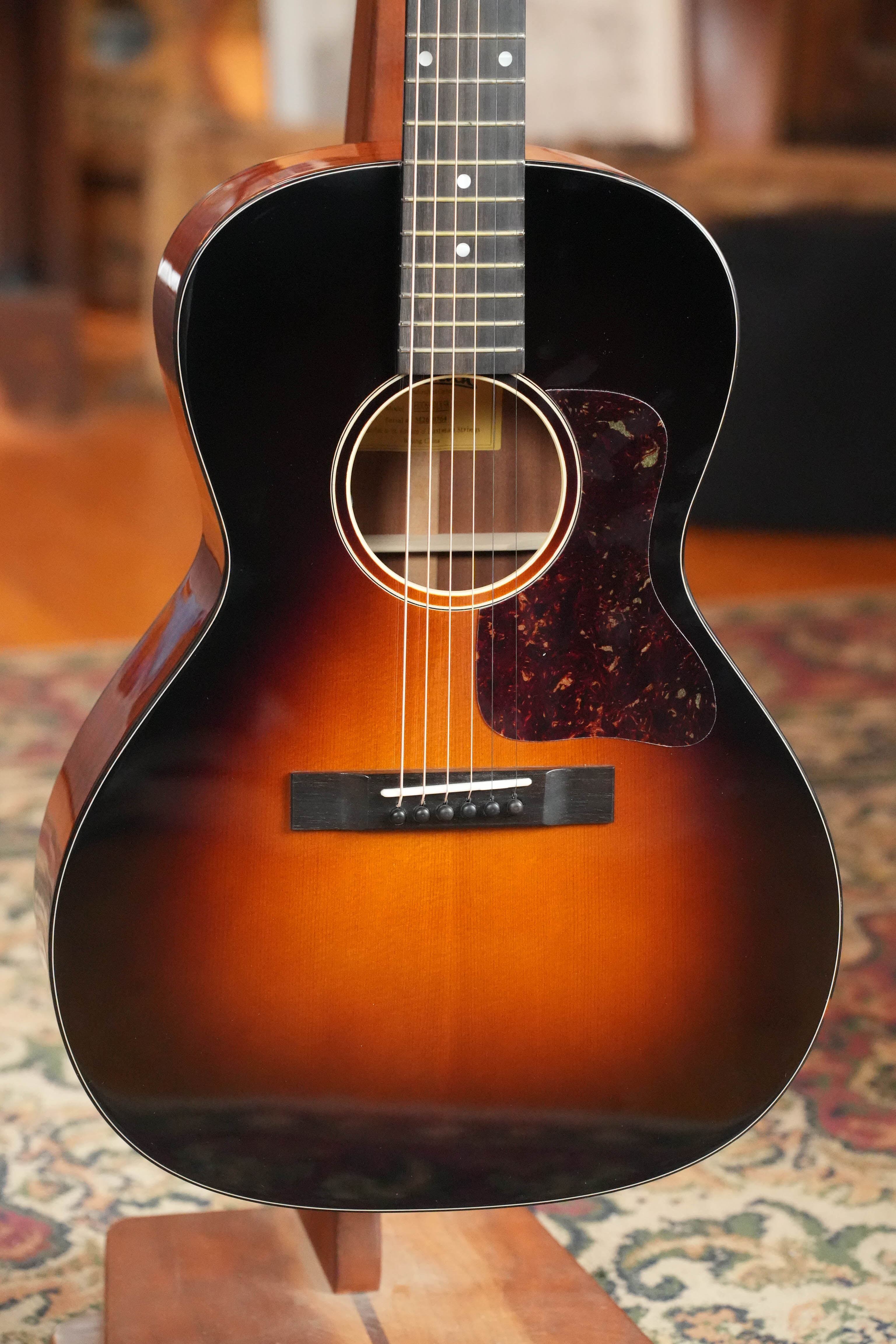 The Eastman Guitars E1OOSS-DLX-SB Sunburst 00 Acoustic-Electric Guitar #1764, featuring a Truetone sunburst finish and round soundhole, stands on a wooden stand indoors on a patterned rug.