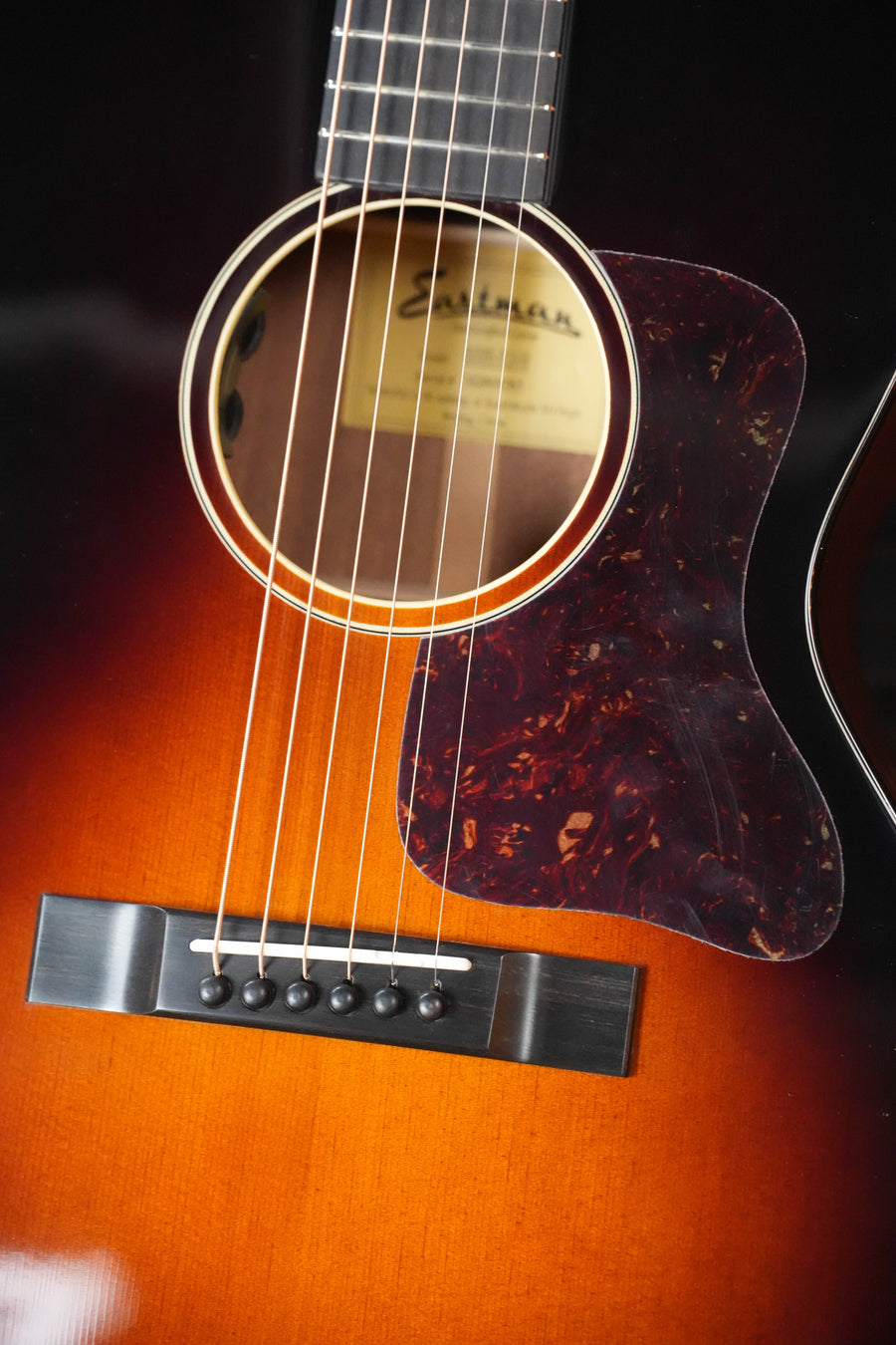Close-up of the Eastman E1OOSS-DLX-SB Sunburst 00 Acoustic-Electric Guitar by Eastman Guitars, showing its glossy Truetone sunburst finish, strings, bridge, pickguard, sound hole with label inside, and a sleek mahogany neck.