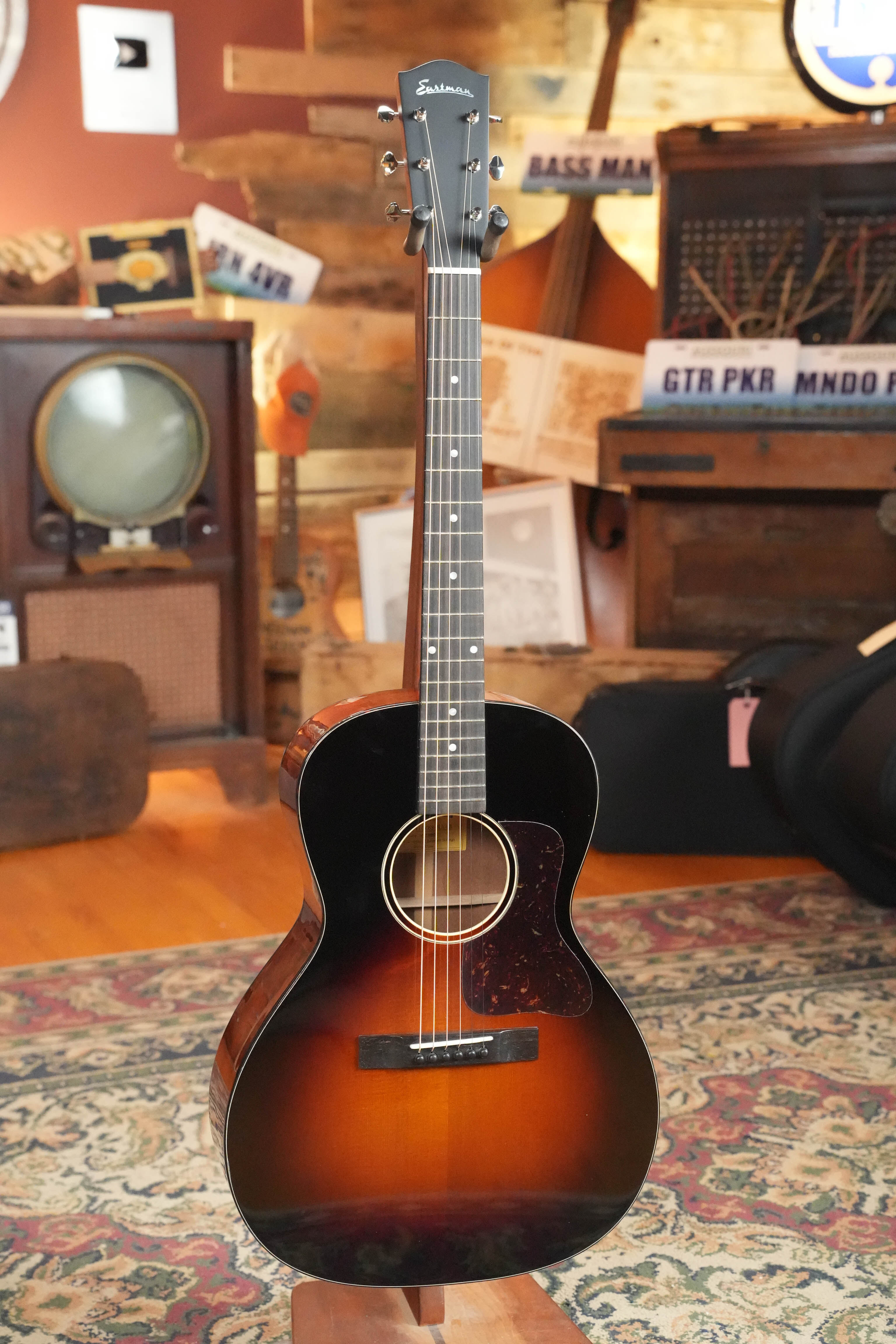 The Eastman Guitars E1OOSS-DLX-SB Sunburst 00 Acoustic-Electric Guitar #8804 rests on a patterned rug in a cozy, vintage-inspired room with an old TV, bass, and music-themed signs evoking a pre-war atmosphere.