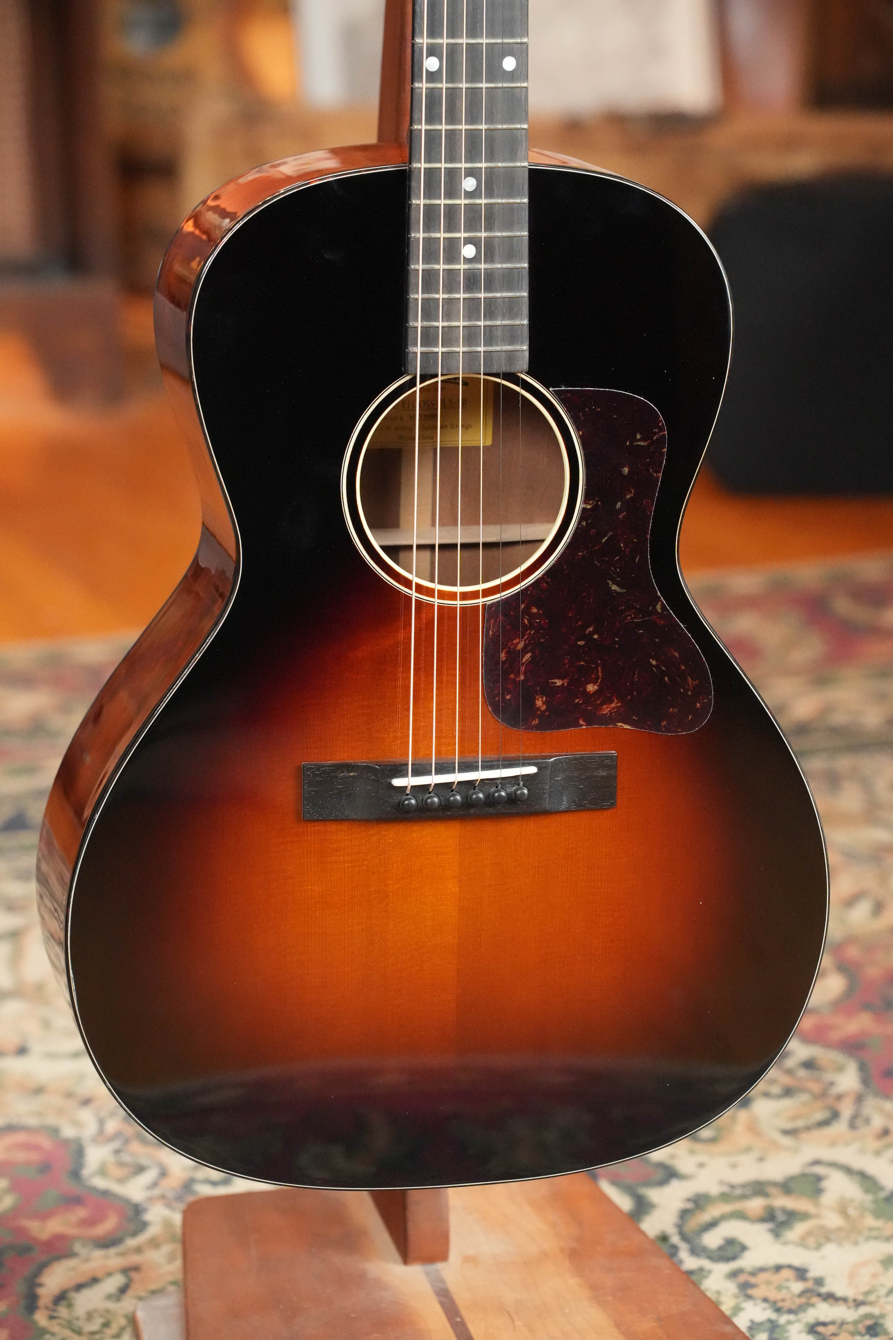 The Eastman Guitars E1OOSS-DLX-SB Sunburst 00 Acoustic-Electric Guitar #8804, featuring a Truetone sunburst finish and tortoiseshell-style pickguard, stands on a wooden stand over a patterned rug, radiating vintage pre-war charm.