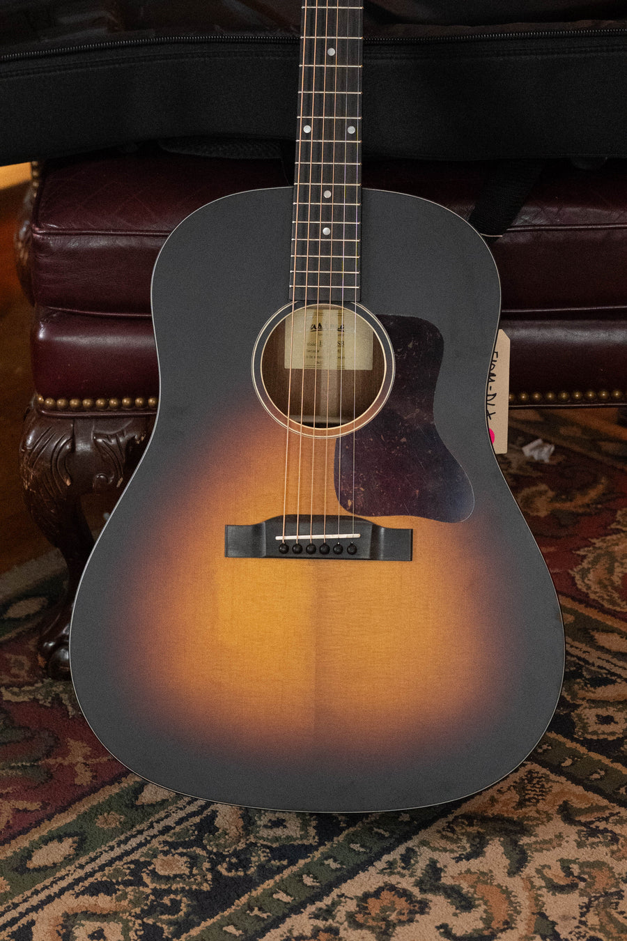 An Eastman Guitars E1SS-SB Sitka/Sapele Sunburst Slope Shoulder Dreadnought Acoustic Guitar #1441 stands upright on an ornate, dark leather chair with carved wooden legs, its sunburst finish displayed atop a patterned rug.