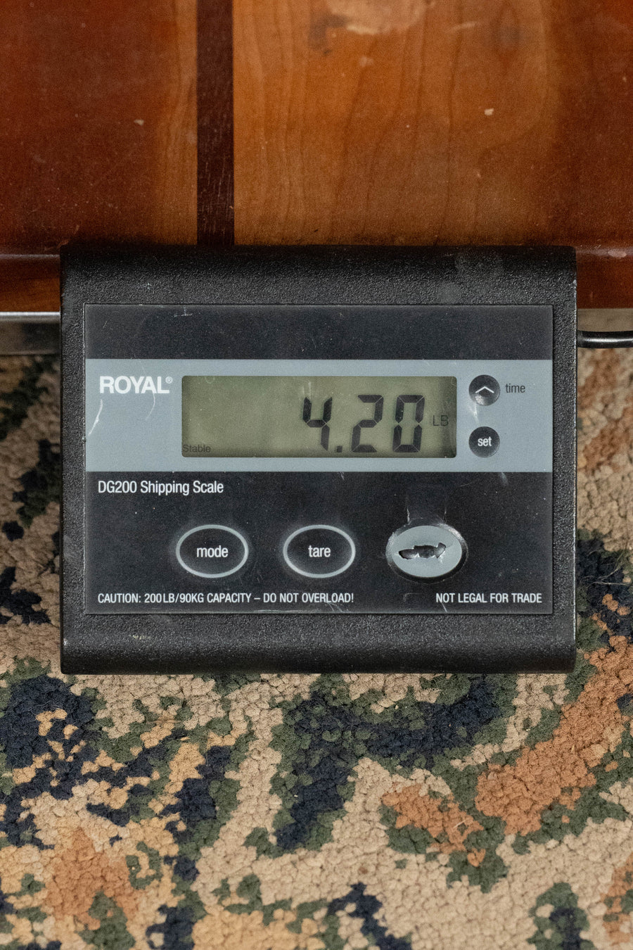 A digital shipping scale on a patterned carpet shows 4.20 lbs, likely weighing an Eastman Guitars E1SS-SB Sitka/Sapele Sunburst Slope Shoulder Dreadnought Acoustic Guitar #1441. The black scale features labeled buttons and cautions about its 200-lb capacity.