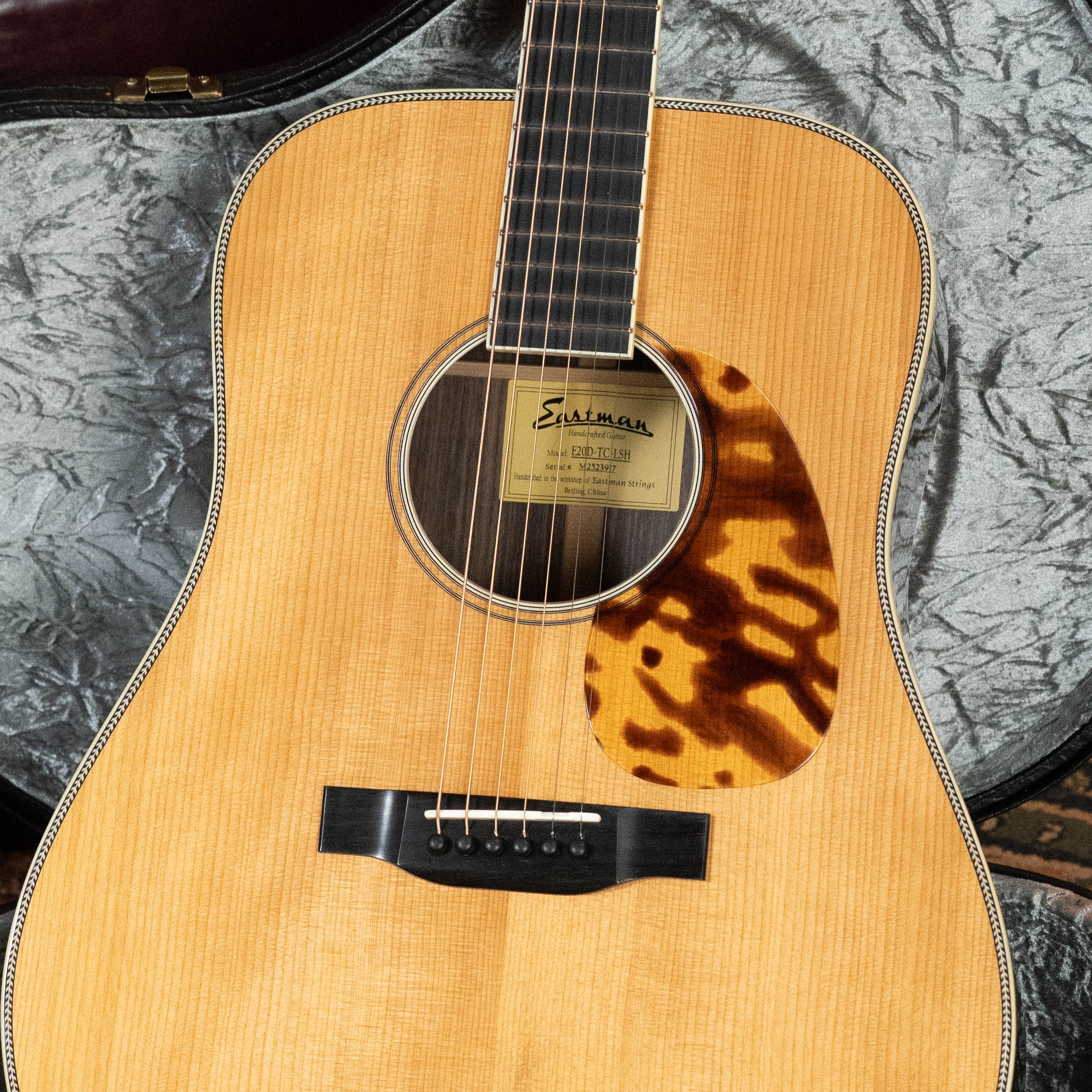 A close-up of the Eastman Guitars E20D-LSH-TC #3917 Large Soundhole Dreadnought, featuring a thermo-cured Adirondack spruce top, light wood finish, tortoiseshell pickguard, and pearl inlay, resting in an open gray-lined hard case.