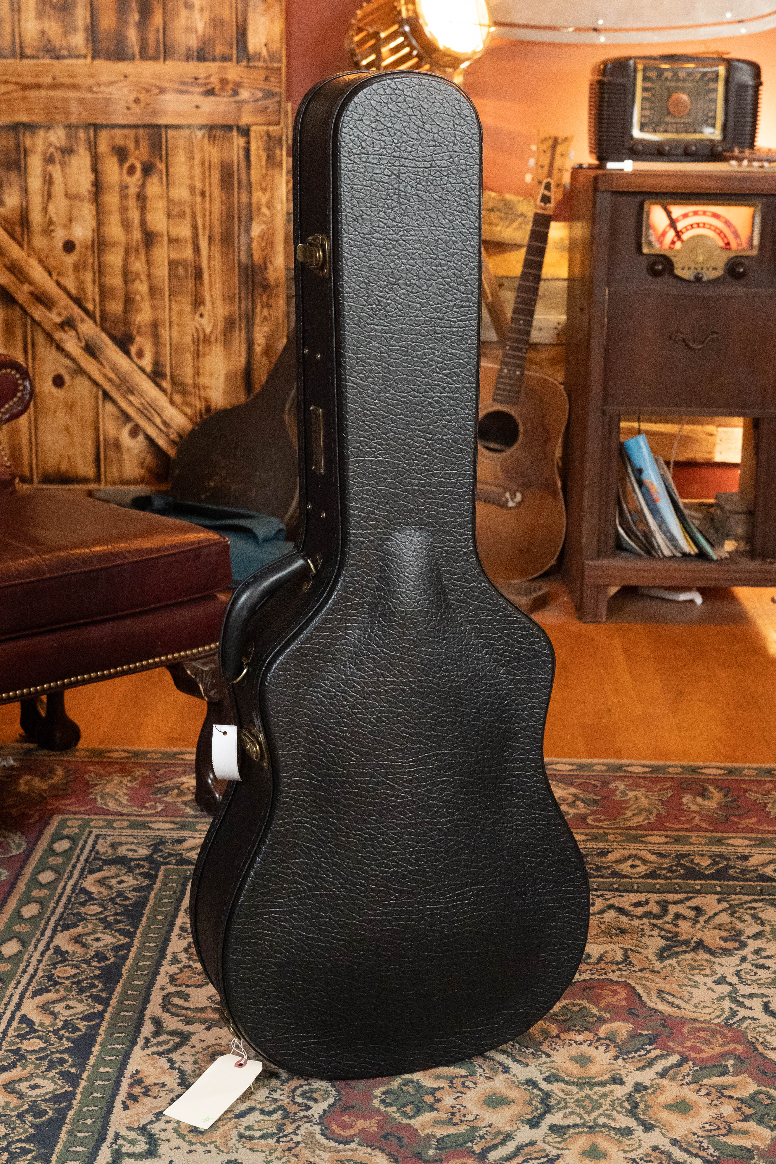 A black hard-shell case stands on a patterned rug beside an Eastman Guitars E20D-LSH-TC Thermo-Cured Adirondack/Rosewood Large Soundhole Dreadnought Acoustic Guitar #3917, with books, a vintage radio, and rustic decor in the cozy room background.