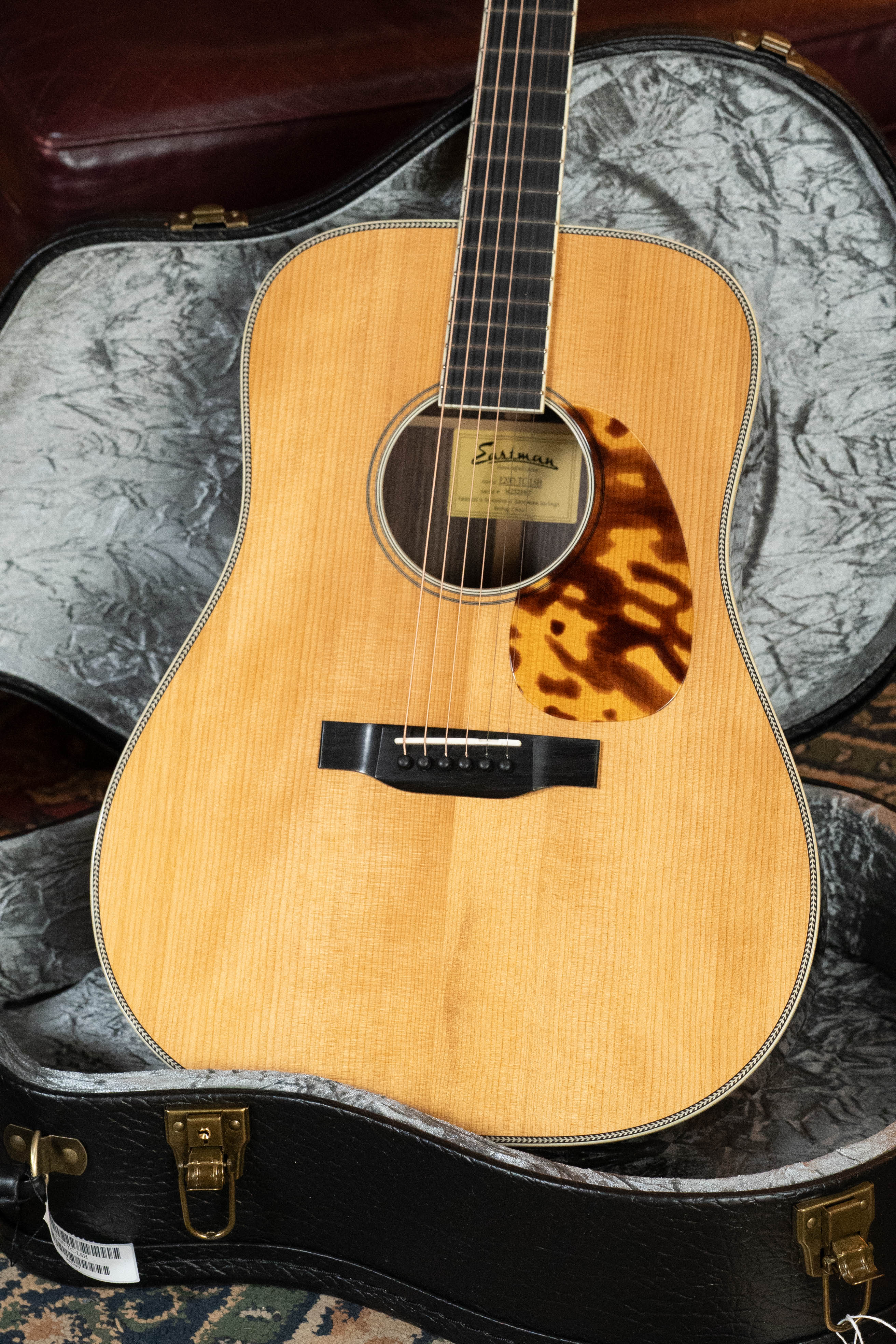 An Eastman Guitars E20D-LSH-TC #3917 acoustic, featuring a Thermo-Cured Adirondack Spruce top and rosewood body, with a tortoiseshell pickguard, rests in an open black padded case on a patterned rug.