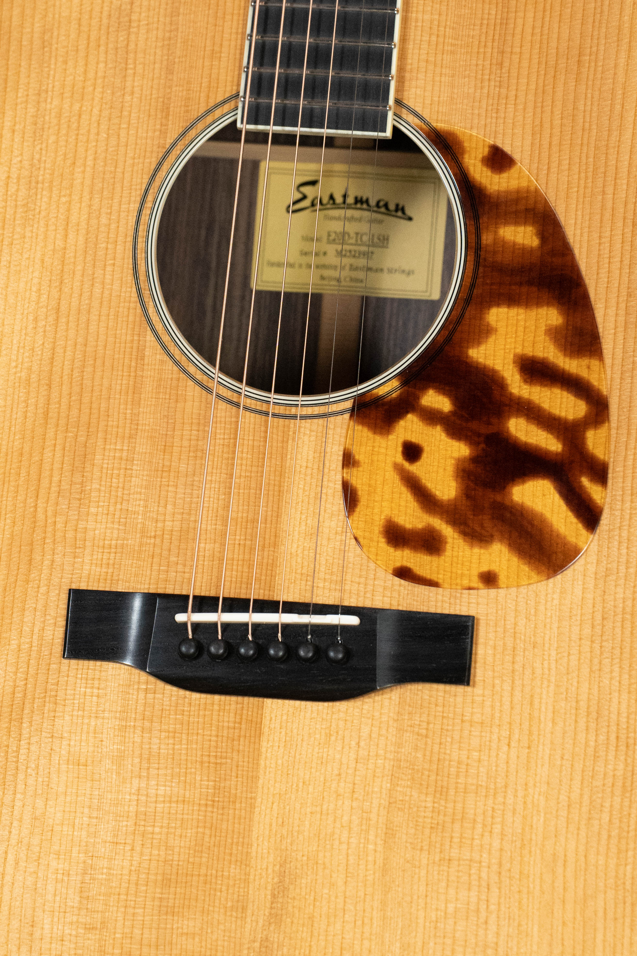 Close-up of the Eastman Guitars E20D-LSH-TC #3917 dreadnought, highlighting its bridge, strings, tortoiseshell-style pickguard, and large soundhole label. Crafted with thermo-cured Adirondack spruce for enhanced tone.