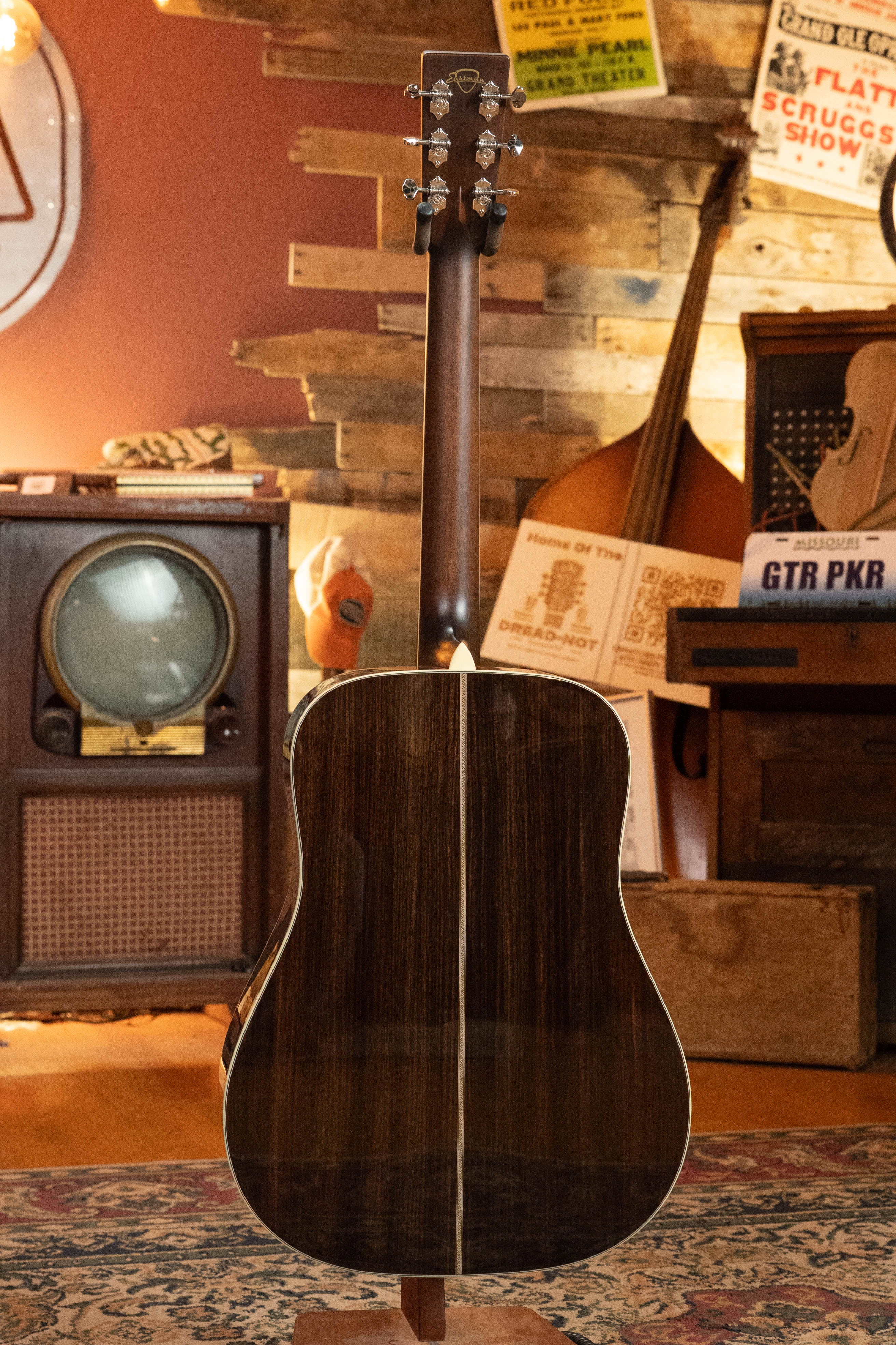 An Eastman Guitars E20D-LSH-TC Thermo-Cured Adirondack/Rosewood Large Soundhole Dreadnought Acoustic Guitar (#3917) stands upright in a cozy vintage room with a patterned rug, old TV, wood-paneled walls, posters, and other instruments.