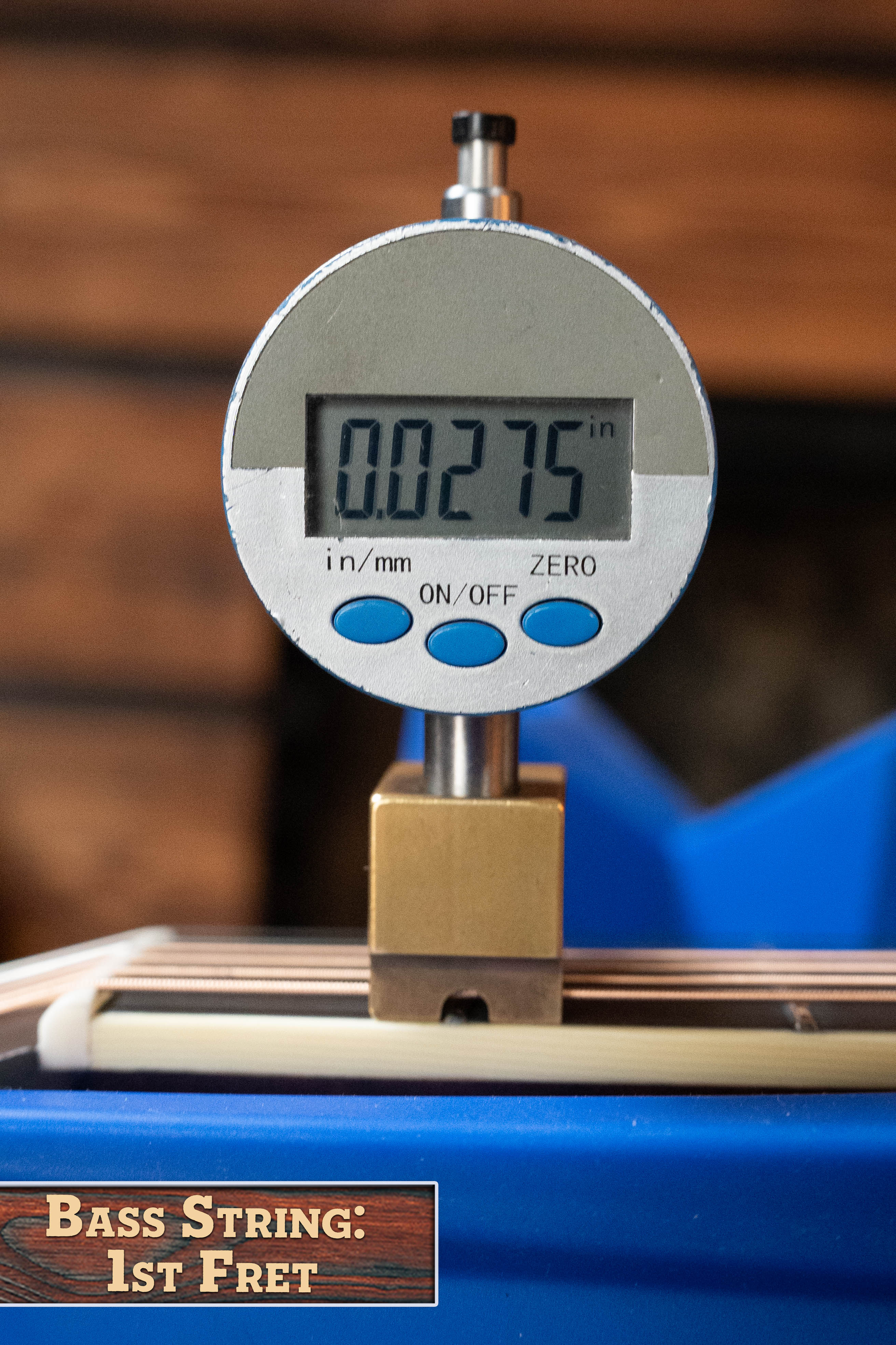 A digital caliper shows 0.0275 inches above the first fret of an Eastman E20D-LSH-TC Thermo-Cured Adirondack/Rosewood Dreadnought by Eastman Guitars, with blurred wood paneling suggesting an acoustic guitar workshop. Label: Bass String 1st Fret.