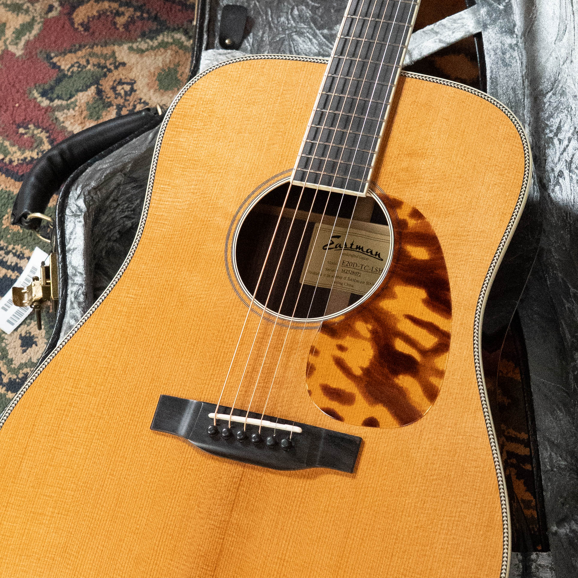 An Eastman Guitars E20D-LSH-TC Thermo-Cured Adirondack/Rosewood Large Soundhole Dreadnought Acoustic Guitar (#8072) with tortoiseshell pickguard rests in an open hard case on a patterned carpet.