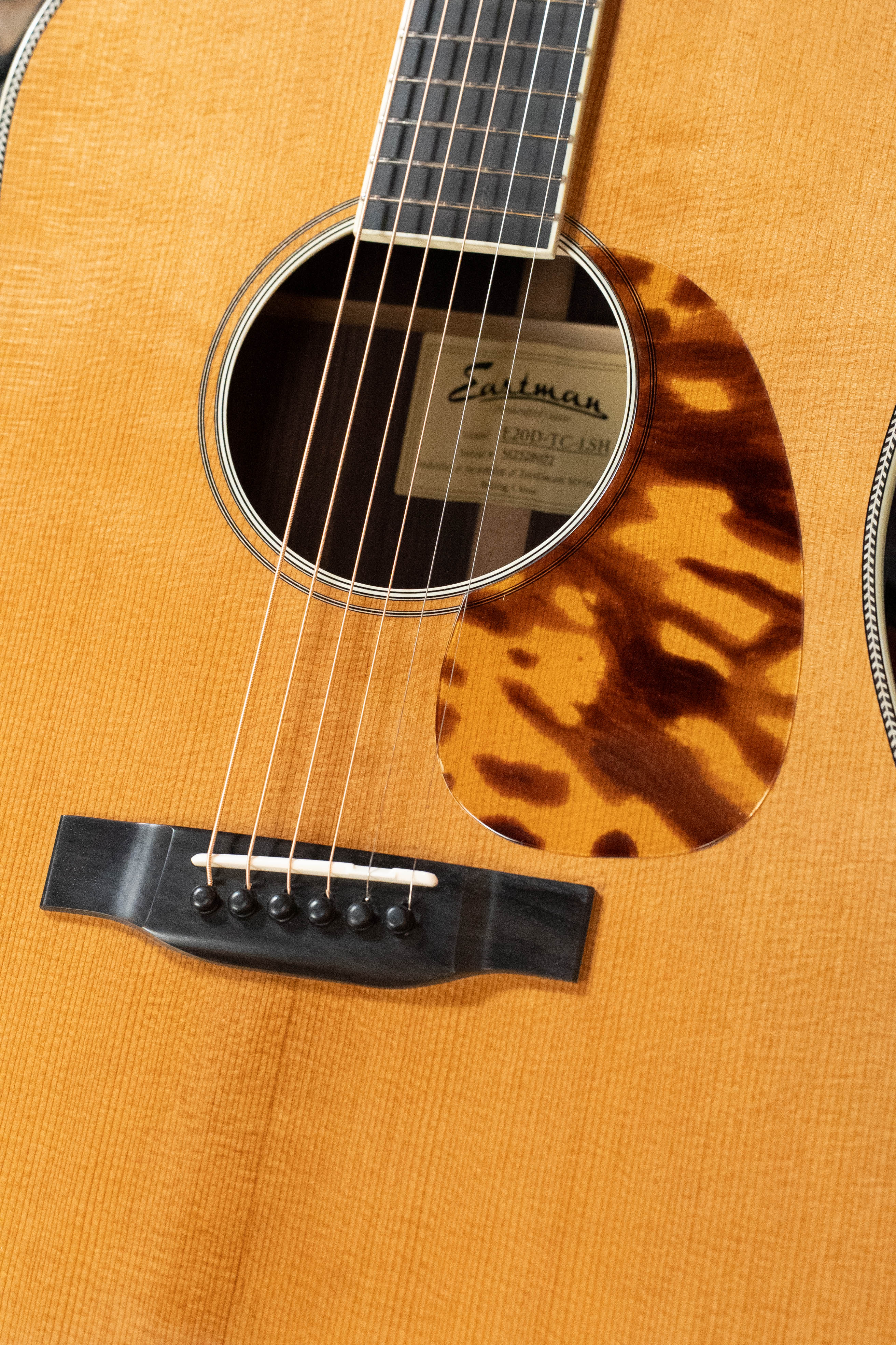 Close-up of the Eastman Guitars E20D-LSH-TC #8072 large soundhole dreadnought, showing strings, bridge, tortoiseshell pickguard, and label; features premium thermo-cured Adirondack Spruce top for enhanced tone.