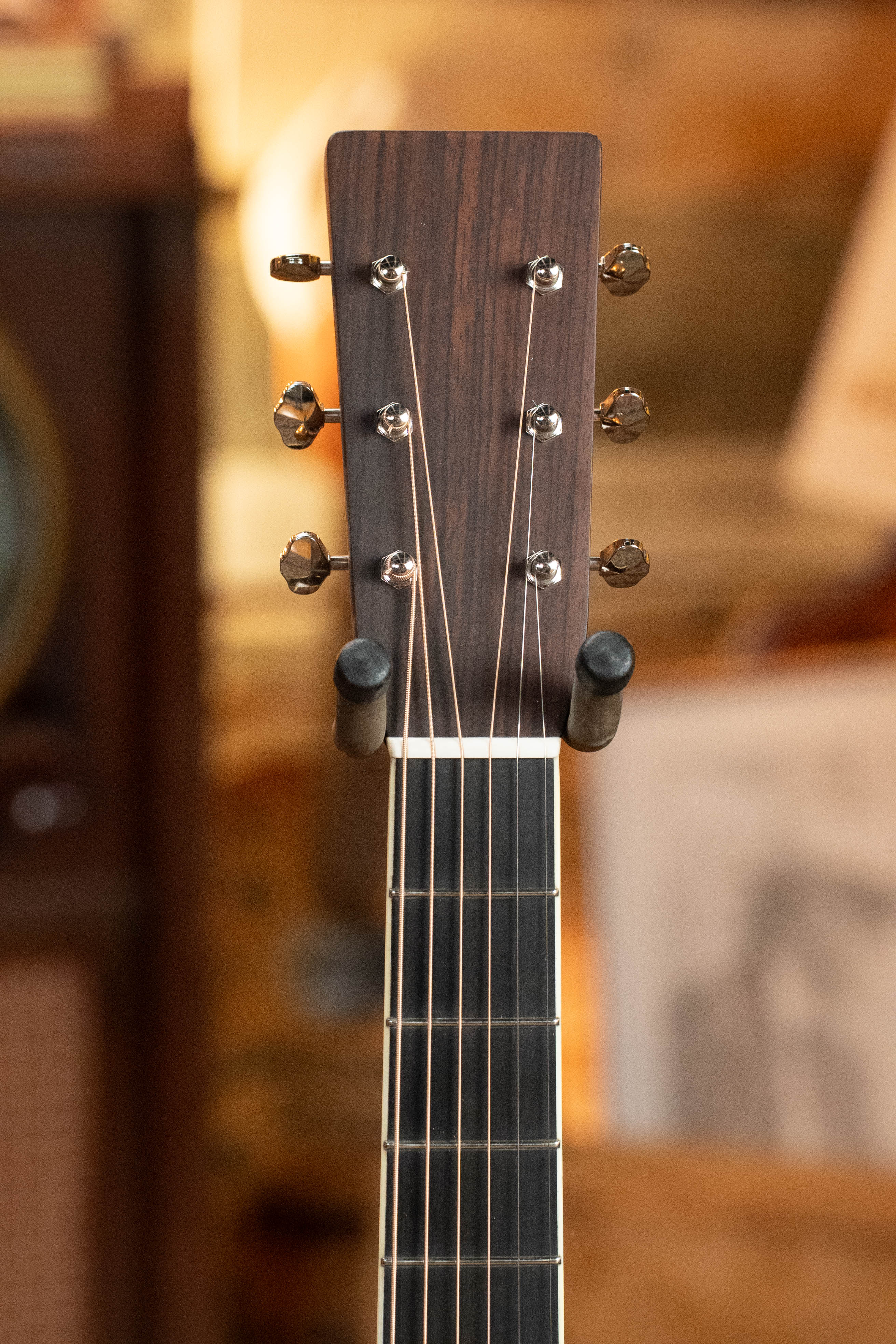Close-up of the Eastman Guitars E20D-LSH-TC #8072 headstock with six tuning pegs, set against a blurred warm background. This dreadnought features thermo-cured Adirondack spruce and rosewood, displayed upright on a stand.