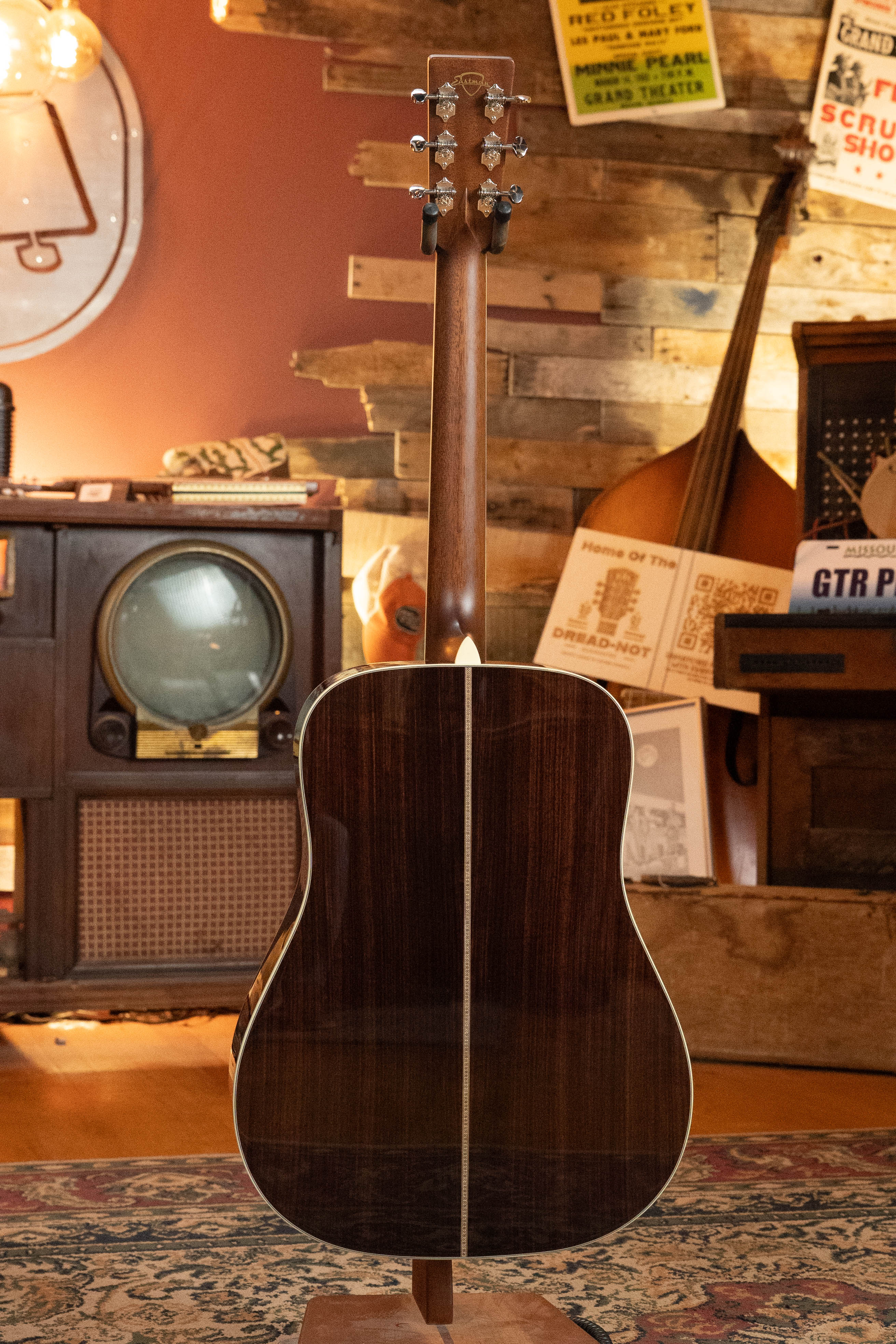 A rear view of the Eastman Guitars E20D-LSH-TC Thermo-Cured Adirondack/Rosewood Dreadnought #8072 stands on a wooden stand in a cozy room with vintage electronics, music posters, and rustic wooden walls.