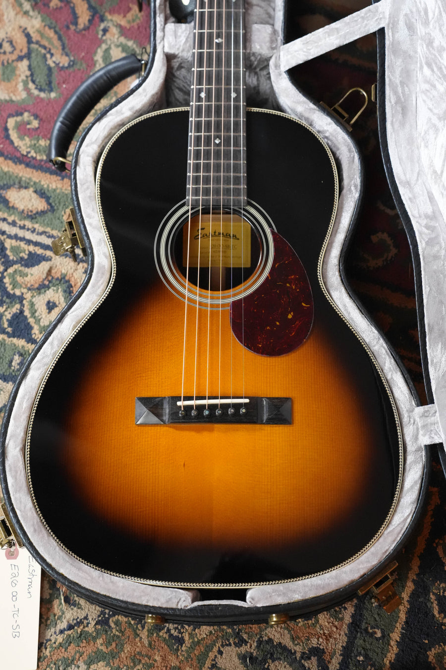 An Eastman Guitars E20OO-TC-SB Thermo-Cured Adirondack/Rosewood Sunburst OO Acoustic Guitar #8110 with a tortoiseshell pickguard sits in a gray-lined hard case on a patterned rug, a handwritten price tag visible at the bottom left.