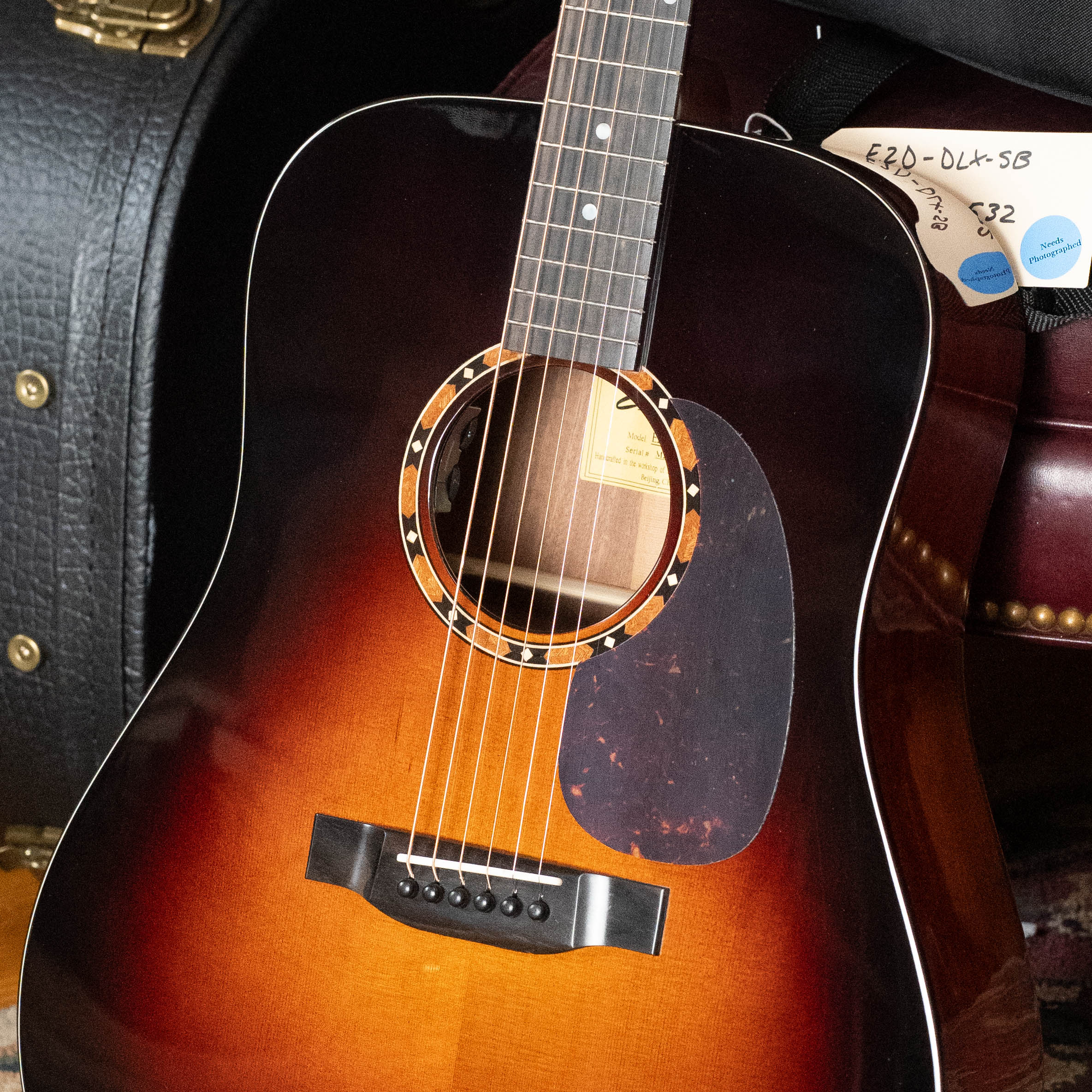 A close-up of the Eastman Guitars E2D-DLX-SB Cedar/Sapele Sunburst Dreadnought Guitar w/Fishman Pickup #1532 shows the strings, soundhole, pickguard, neck, a black case, and tags in the background.